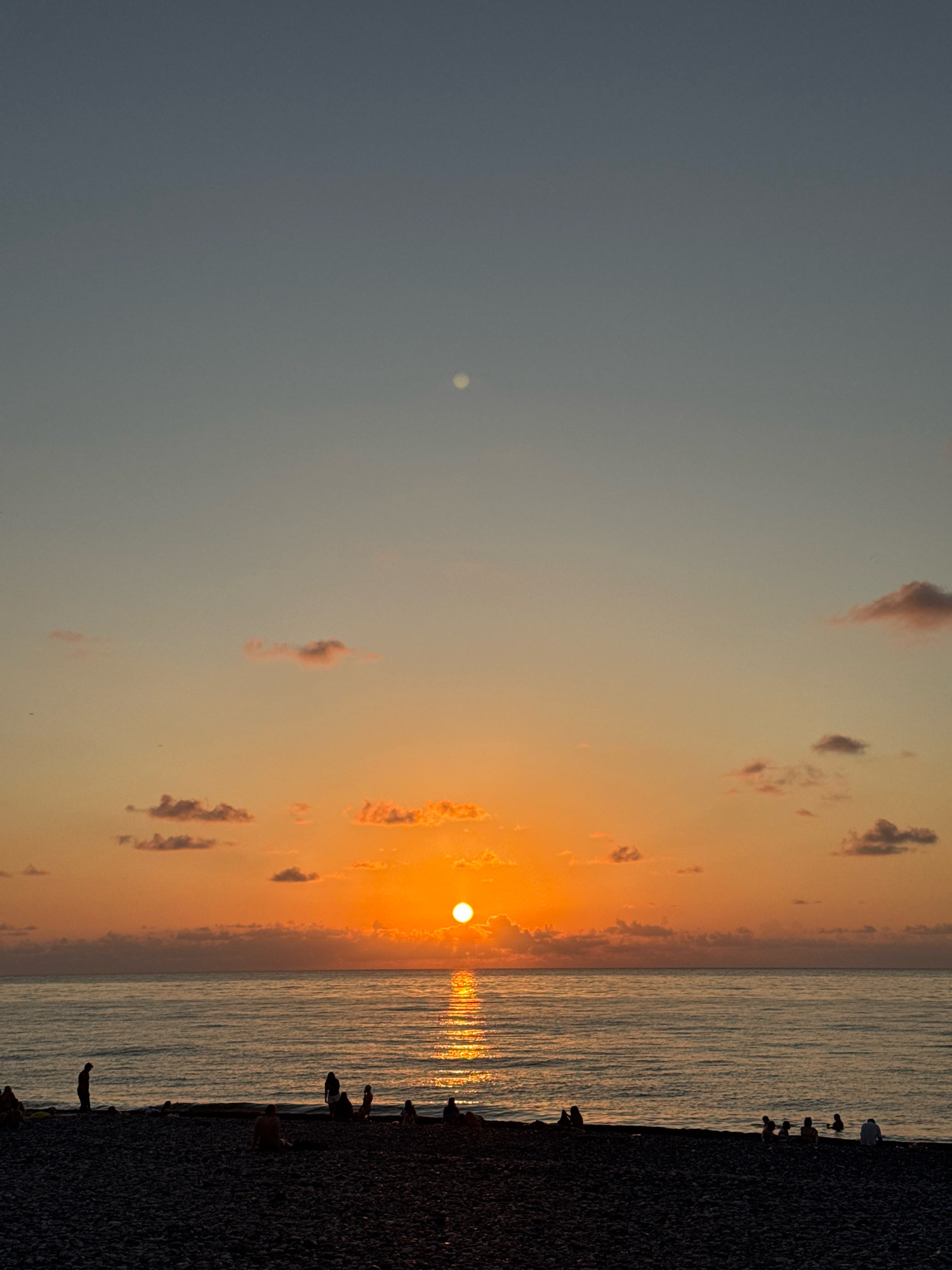 This is a photograph of a sunset over the Black Sea, taken from a pebble beach in Batumi, Georgia. The sun is a bright orange orb on the horizon, casting a golden reflection on the calm water. The sky above transitions from a pale blue to hues of orange and pink, with a few scattered clouds. Silhouettes of people can be seen on the beach, some standing and others sitting, enjoying the view. The waves gently lap at the shore, and the overall atmosphere is serene.