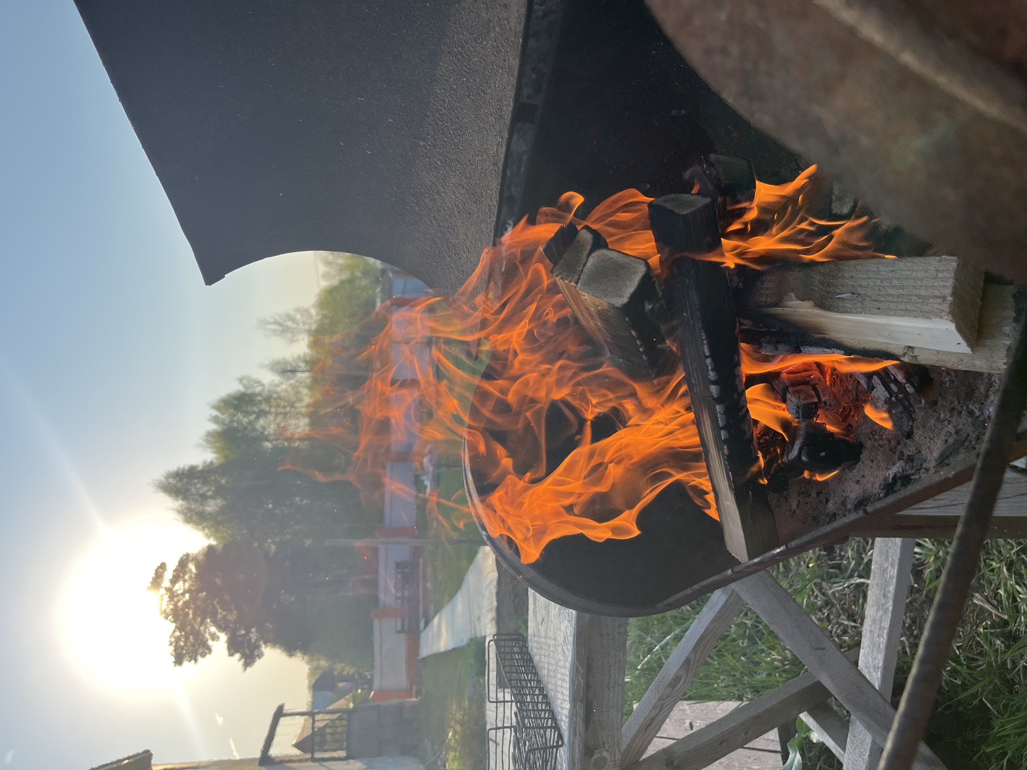 Here's a description of the image:
The image captures a close-up, slightly rotated view of a fire burning inside a grill. The fire is intense, with large, bright orange flames engulfing pieces of wood. The grill appears to be made of dark metal and sits on a wooden frame.
In the background, partially visible through the smoke and grill structure, is a sunlit outdoor scene. There are trees and a walkway. In the distance, there are houses with orange roof.
Although the image might be taken somewhere near Chaikovsky, Russia, based on certain features like the architecture of the house visible in the background, there is insufficient evidence to conclusively confirm its location within the city.