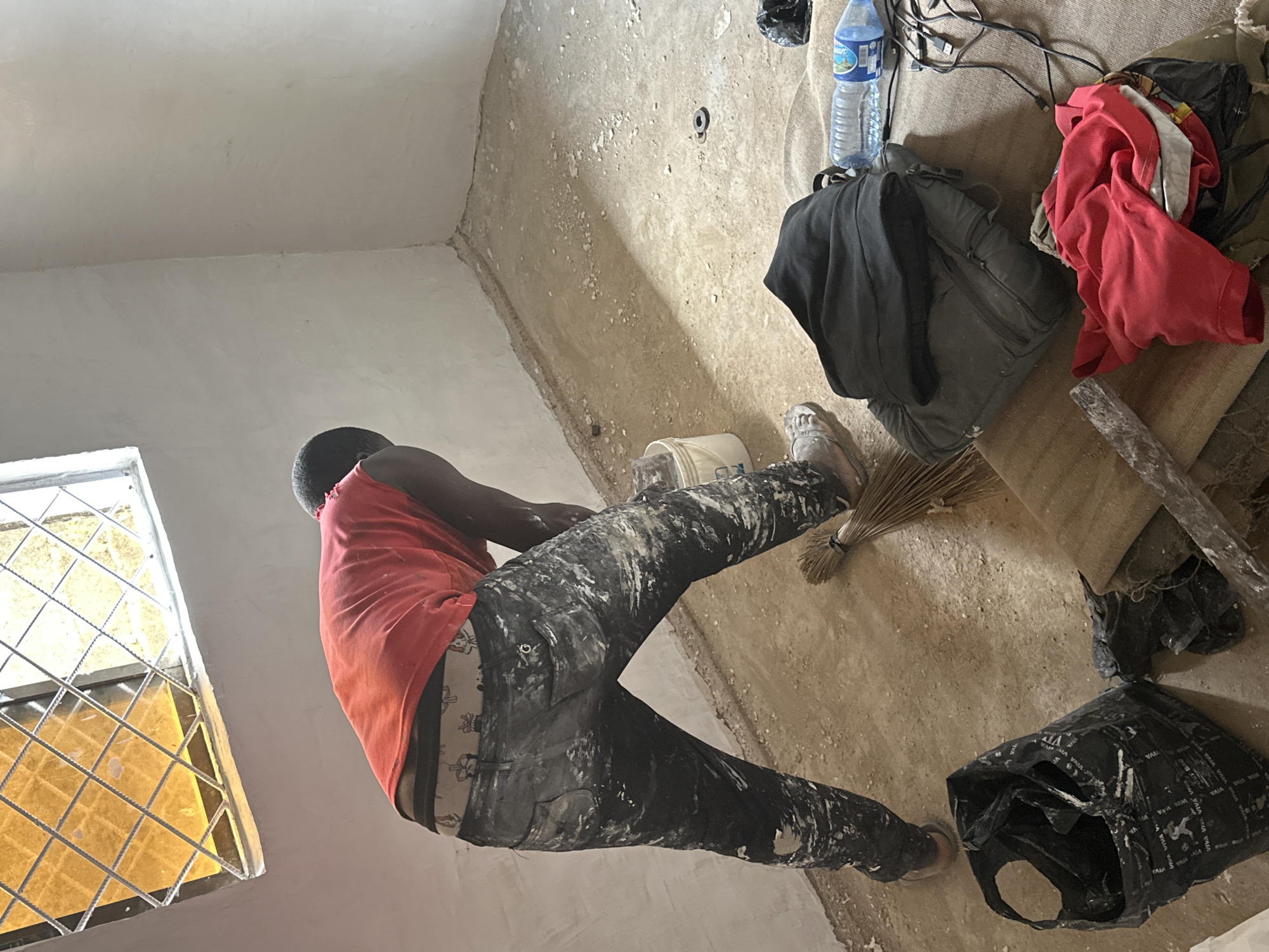 This image captures an indoor scene in Bukuru, Nigeria, depicting a man at work in what appears to be a room undergoing renovation or construction. The man, viewed from the back and side, has a dark complexion and is wearing a red tank top and dark trousers heavily stained with white paint or plaster splatters. A patterned boxer waistband is visible above his pants. His light-colored shoes or sandals are also covered in white residue.
He is bent over, facing a light-colored wall, and is holding a white bucket, suggesting he is engaged in plastering or painting. The walls are a light grey or off-white, with some areas appearing freshly done while a corner shows rougher, unplastered surfaces and minor cracks. The floor is a rough, dusty concrete surface, scattered with white debris and small stones.
To the right of the man, various items are resting on the floor, including a dark grey backpack, a crumpled red shirt, a plastic water bottle, and tangled black wires, possibly USB cables. A small, natural-fiber broom lies near his feet. Further right, a black plastic bag, also covered in white stains, and a wooden plank are visible. The water bottle and the black plastic bag both display the text "VIVA".
On the left side of the frame, a window with a metal grid allows bright daylight into the room, illuminating its yellow-tinted panes. The overall scene conveys an active work environment in an unfinished or utilitarian space.