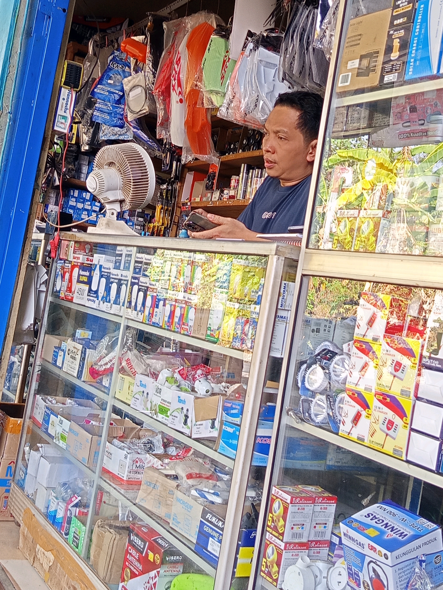The image depicts the interior of a small shop, likely a hardware or electronics store, situated in the city of Karang Sari, Indonesia. The shop is filled with various goods, displayed on shelves and in glass cabinets. Prominently featured are electrical components, such as light bulbs, chargers, and fans, alongside what appear to be motorcycle parts, including orange fenders.
In the foreground, a man with short black hair is looking to his left, holding a smartphone in his right hand. He is wearing a dark blue t-shirt with a white logo. He is positioned behind a glass counter, through which numerous products are visible.
The shop's interior is densely packed with merchandise. On the shelves above the man are various packaged goods, including boxes and bags containing parts. In front of the man, the glass display cases showcase more products, sorted by type and brand. Some notable items include "Hannochs Supra" light bulbs, "Morgen" boxes, "Vord Torch" products, and "Winngas" items, suggesting a range of electrical and gas-related supplies.
The overall environment appears to be that of a typical Indonesian neighborhood store, characterized by its crowded display of goods and a direct, no-frills approach to retail. The lighting suggests it is daytime, with natural light filtering into the shop from the front entrance. The blue framing of the shop's entrance is visible on the left side of the image.
