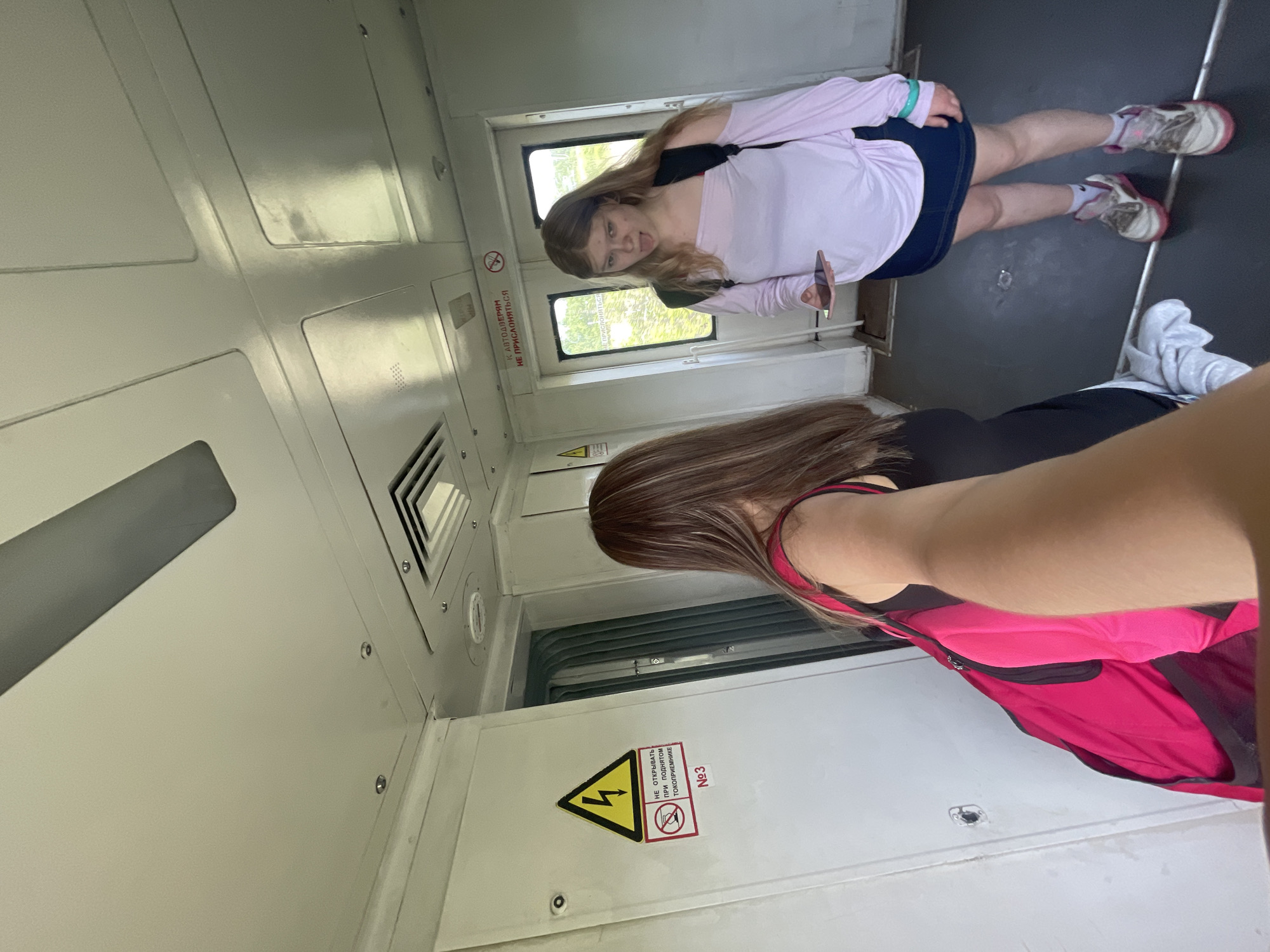This image depicts the interior of a passenger train car, consistent with a location in Russia, such as Priozersk. The perspective is a wide-angle, selfie-style photograph.
In the immediate foreground, the right arm and shoulder of an individual taking the photograph are visible, extending from the lower right toward the center. This individual has long, light brown hair and is wearing a black top with a bright pink backpack on the right shoulder.
To the right of the frame, a young female stands facing the camera. She has long, light brown hair, an open mouth with her tongue extended, and is holding a light-colored smartphone horizontally in her left hand. She is dressed in a light pink, off-the-shoulder long-sleeved top, a dark blue denim mini-skirt, white socks, and white sneakers with pink accents. A black backpack is worn on her back, and a light blue bracelet is visible on her left wrist.
The train car's interior features white paneled walls and a dark grey floor with a horizontal metal strip. On the left wall, a yellow triangular warning sign with a black lightning bolt is prominent. Below it, a red circular prohibition sign depicts a hand attempting to open a panel, accompanied by Cyrillic text stating "НЕ ОТКРЫВАТЬ ПРИ ПОДНЯТОМ ТОКОПРИЕМНИКЕ" (DO NOT OPEN WHEN PANTOGRAPH IS RAISED) and the number "№3". Partially visible to the right of this sign are accordion-style doors.
Further right, a set of double doors with windowpanes reveals green foliage outside, indicating the train is either in motion or stopped in a natural setting. Above these doors, another sign in Cyrillic reads "К АВТОДВЕРЯМ НЕ ПРИСЛОНЯТЬСЯ" (DO NOT LEAN ON AUTO DOORS), alongside a red prohibition symbol of a person leaning. The ceiling is white and paneled, featuring several recessed circular fixtures, a square air vent, and a long, grey rectangular fixture. The train car exhibits signs of wear on the walls and floor.