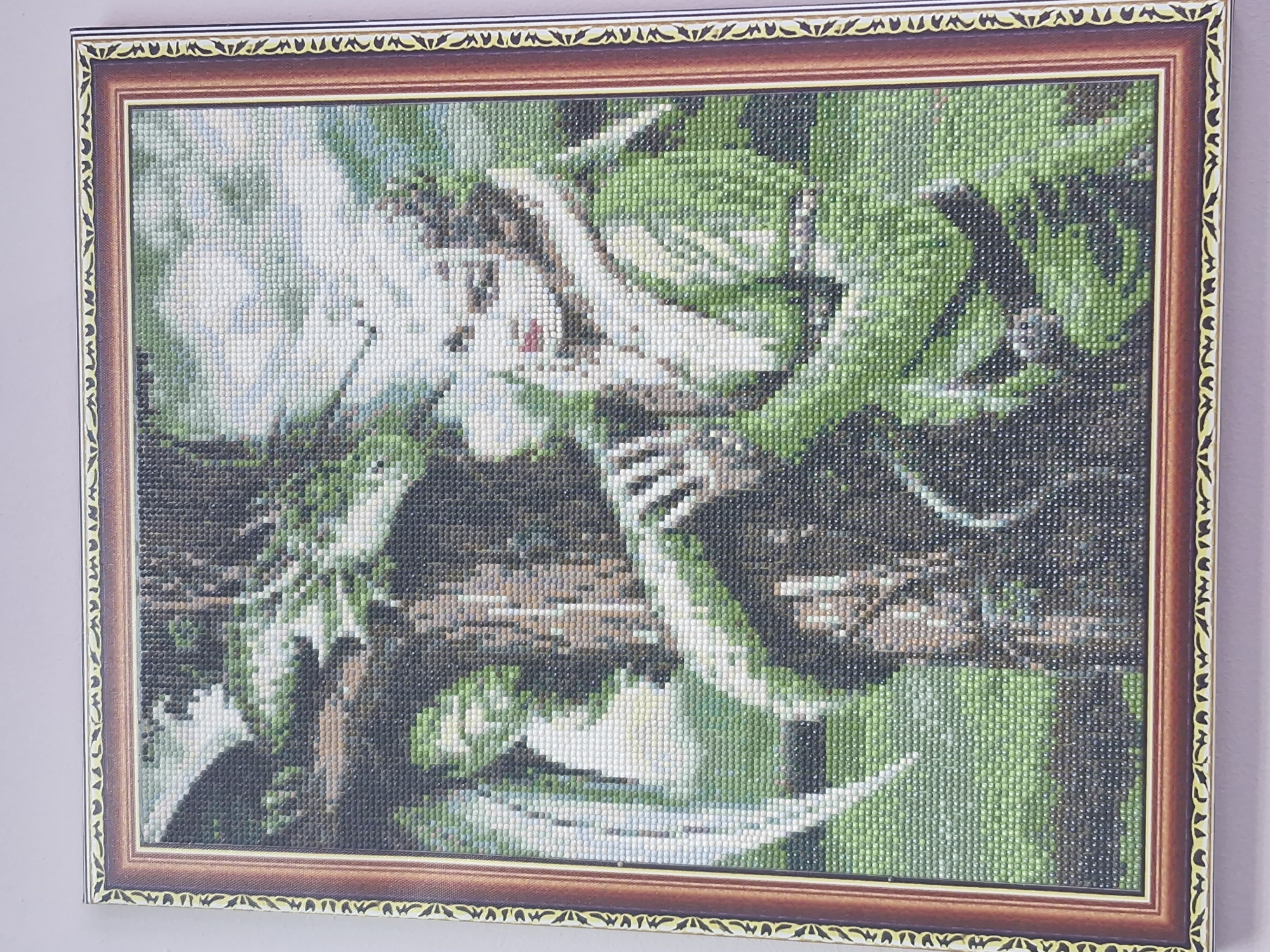 A framed, handcrafted diamond painting depicting a bright green snake winding around a branch with white flowers, possibly orchids, is displayed against a plain, light-colored wall. The painting is intricate, made up of individual diamond beads, and framed in a decorative, two-toned brown frame. The image offers no specific clues to place it in Markovskii, Russia, or to provide any information about people or activities related to the location.