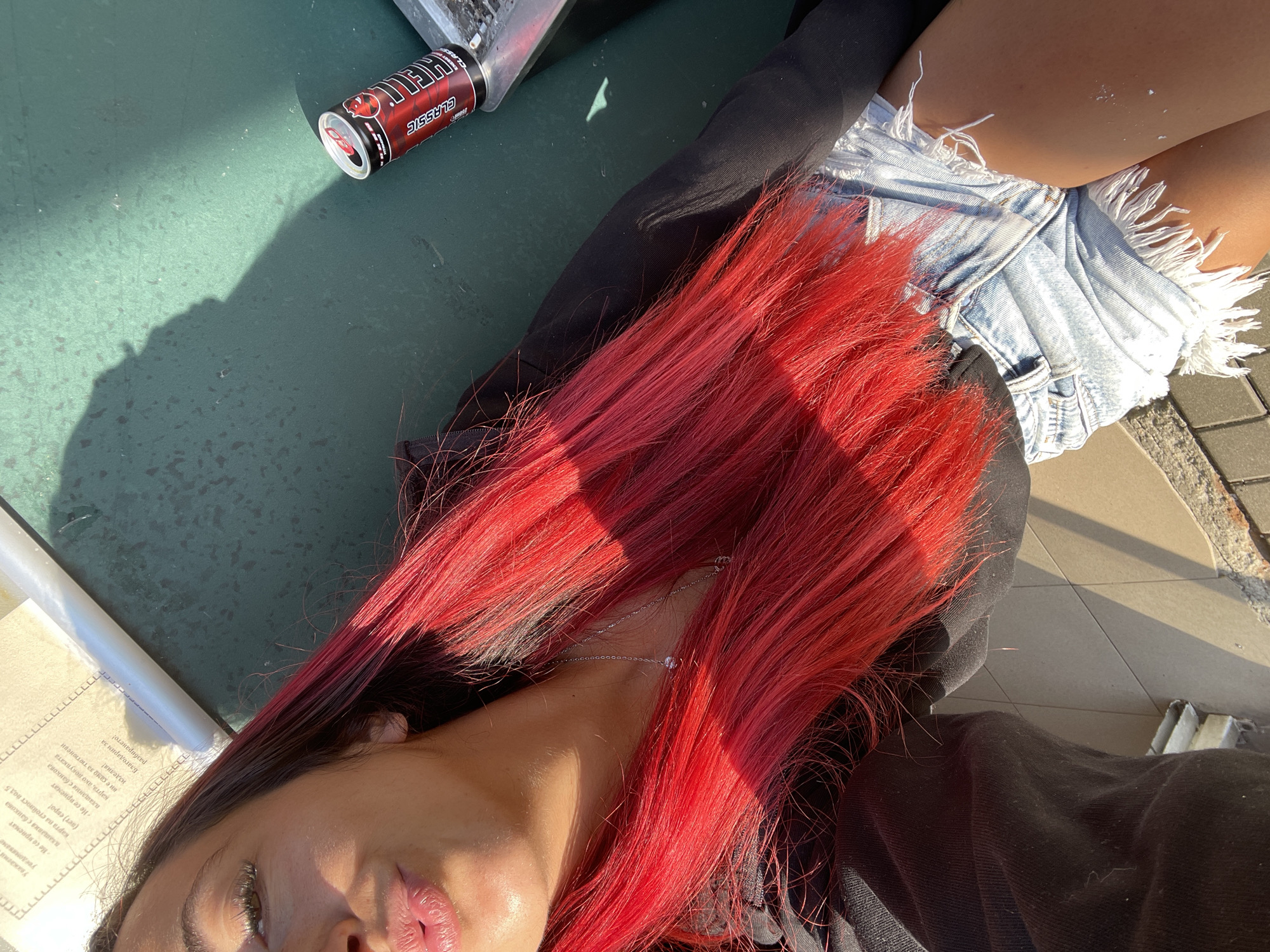 A close-up selfie shows a woman with bright red hair and fair skin. She is wearing denim shorts and a black top. Her lips are pursed, and she is looking towards the camera. Sunlight illuminates her face and hair, casting a shadow on the green surface behind her. A can of "Hell Energy" drink is visible to her right. The background shows a tiled surface and a piece of paper with text in Bulgarian, which appears to be from a menu or informational leaflet, possibly indicating a location in Sofia, Bulgaria, such as a cafe or outdoor seating area.