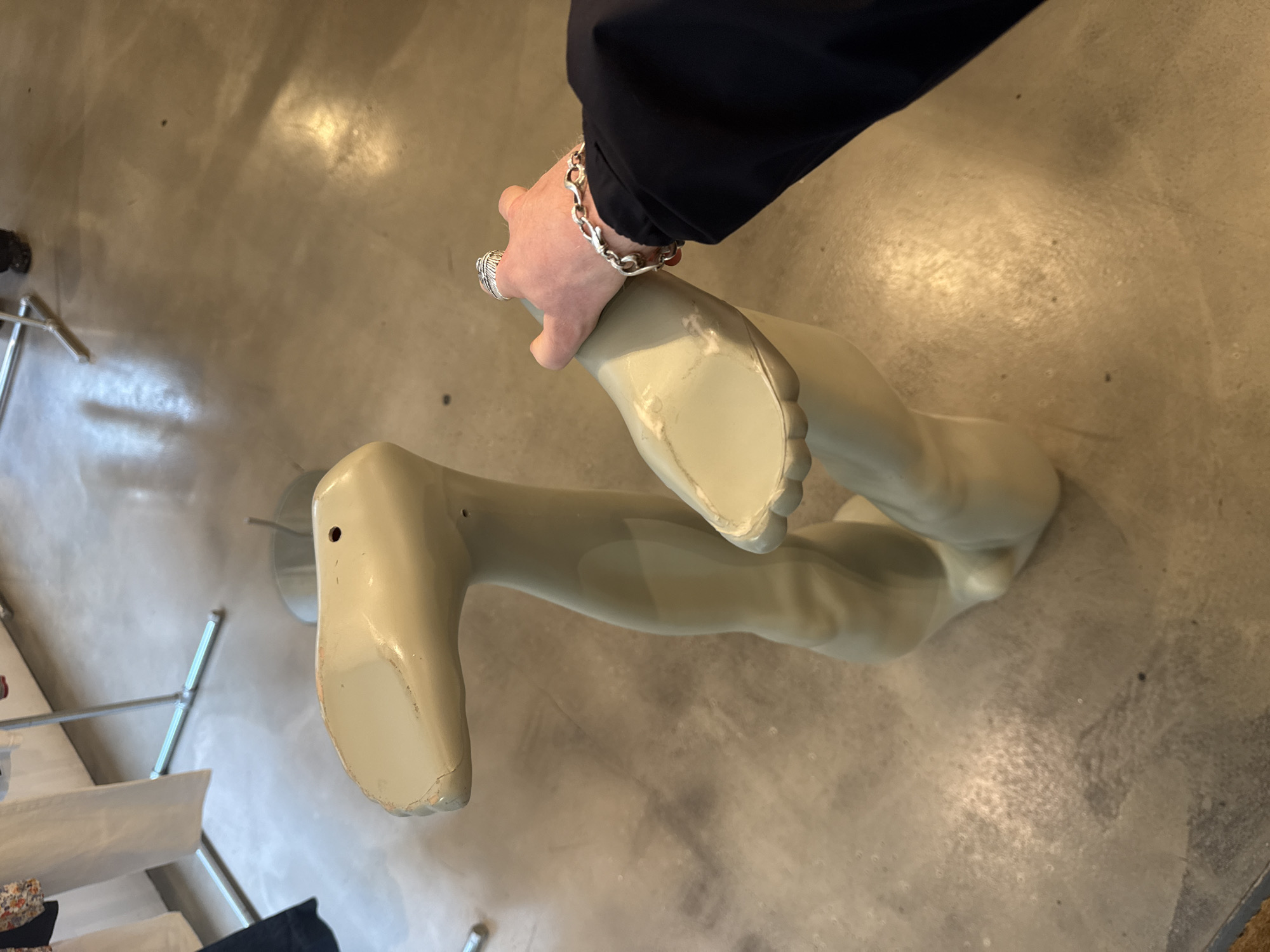 The image depicts a partial view of a mannequin's legs, pale in color, being held up by a person wearing a black sleeve and a silver bracelet and ring. The background shows a gray, possibly concrete floor with a rack visible in the lower-left corner. No information suggests this is taken in London.