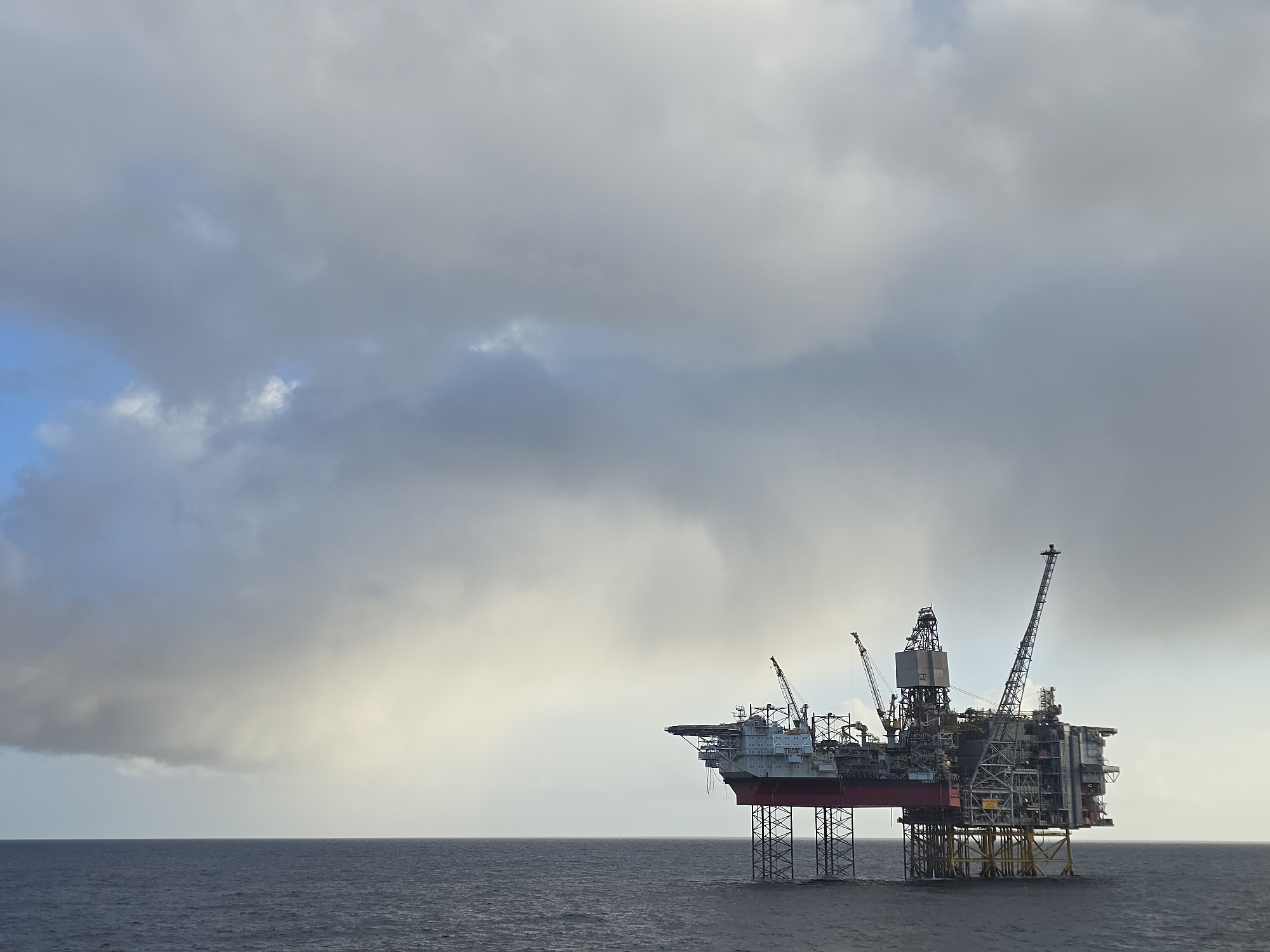 The image depicts an offshore oil platform in the ocean under a cloudy sky. The platform is a complex structure with multiple levels, cranes, and derricks. It appears to be a fixed platform, resting on steel legs anchored to the seabed. The sky is overcast with heavy, gray clouds covering most of the frame. The sea is relatively calm with small waves. The location is Unknown, Unknown.