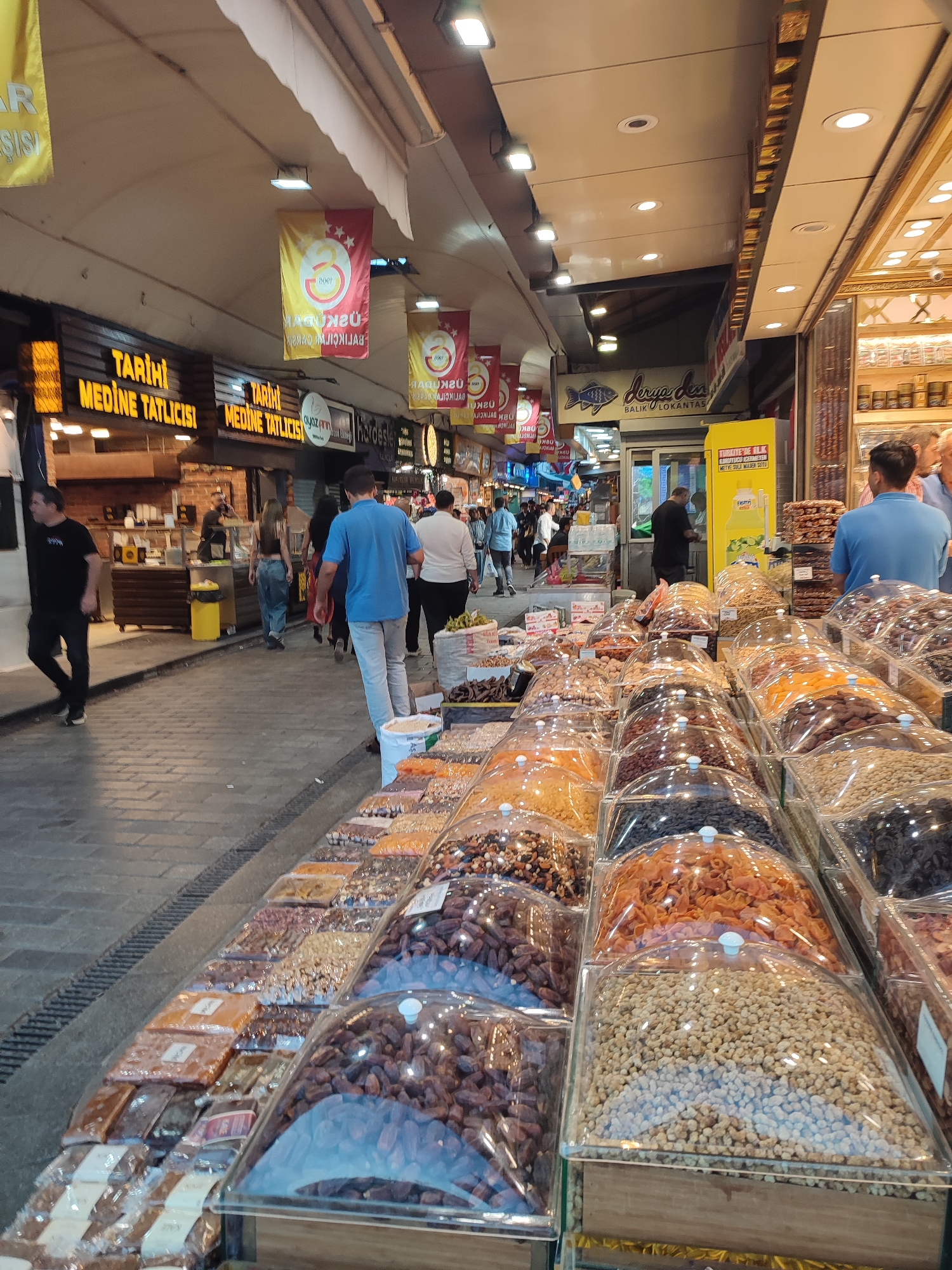 The image depicts a bustling marketplace, likely in Üsküdar, Turkey, given the presence of a Turkish flag banner with the Galatasaray logo and text in Turkish. The scene is characterized by numerous stalls laden with a wide variety of dried fruits, nuts, and sweets, displayed in clear plastic containers and packages.
In the foreground, a vibrant display of goods is presented. Several large, transparent containers showcase an assortment of dried fruits like apricots, dates, and possibly prunes, alongside various nuts and what appear to be candies or confections. The items are meticulously arranged, inviting customers to browse. Behind these stalls, more packaged goods are visible, suggesting a diverse range of products.
The marketplace appears to be an open-air covered walkway, with a cobblestone-like ground. Shops with illuminated signs are visible along the sides. One sign, prominently displayed, reads "Tarihi Medine Tatlicisi," indicating a historic sweet shop. Another sign features a stylized fish logo and the text "Deniz Balik Lokantasi," suggesting a seafood restaurant. Yellow banners with Turkish writing hang overhead.
Several individuals are present in the scene. In the middle ground, a man in a blue t-shirt and grey pants walks away from the camera, while other shoppers navigate the busy thoroughfare. On the left, a man in a black t-shirt and dark pants walks towards the viewer. People are seen interacting with vendors and browsing the merchandise. The overall atmosphere is one of lively commercial activity. The lighting suggests it is daytime, with artificial lights also contributing to the illumination.