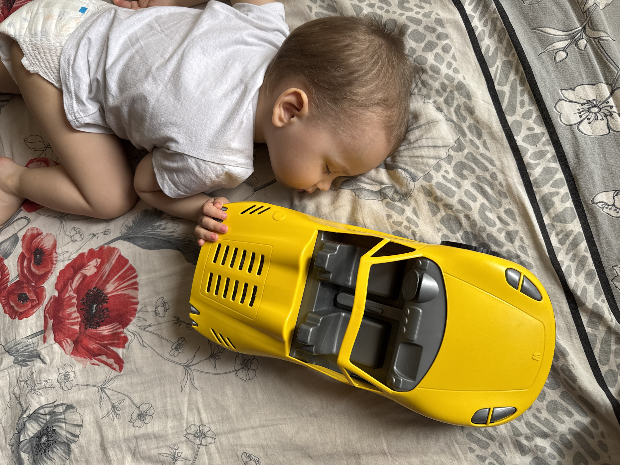 A child sleeps on a patterned bed sheet next to a yellow toy car. The child is wearing a diaper and a white t-shirt. The bed sheet has a floral pattern with red and gray flowers. The toy car is a convertible with a gray interior. It is not possible to determine if this image was taken in Rusanovo, Russia.