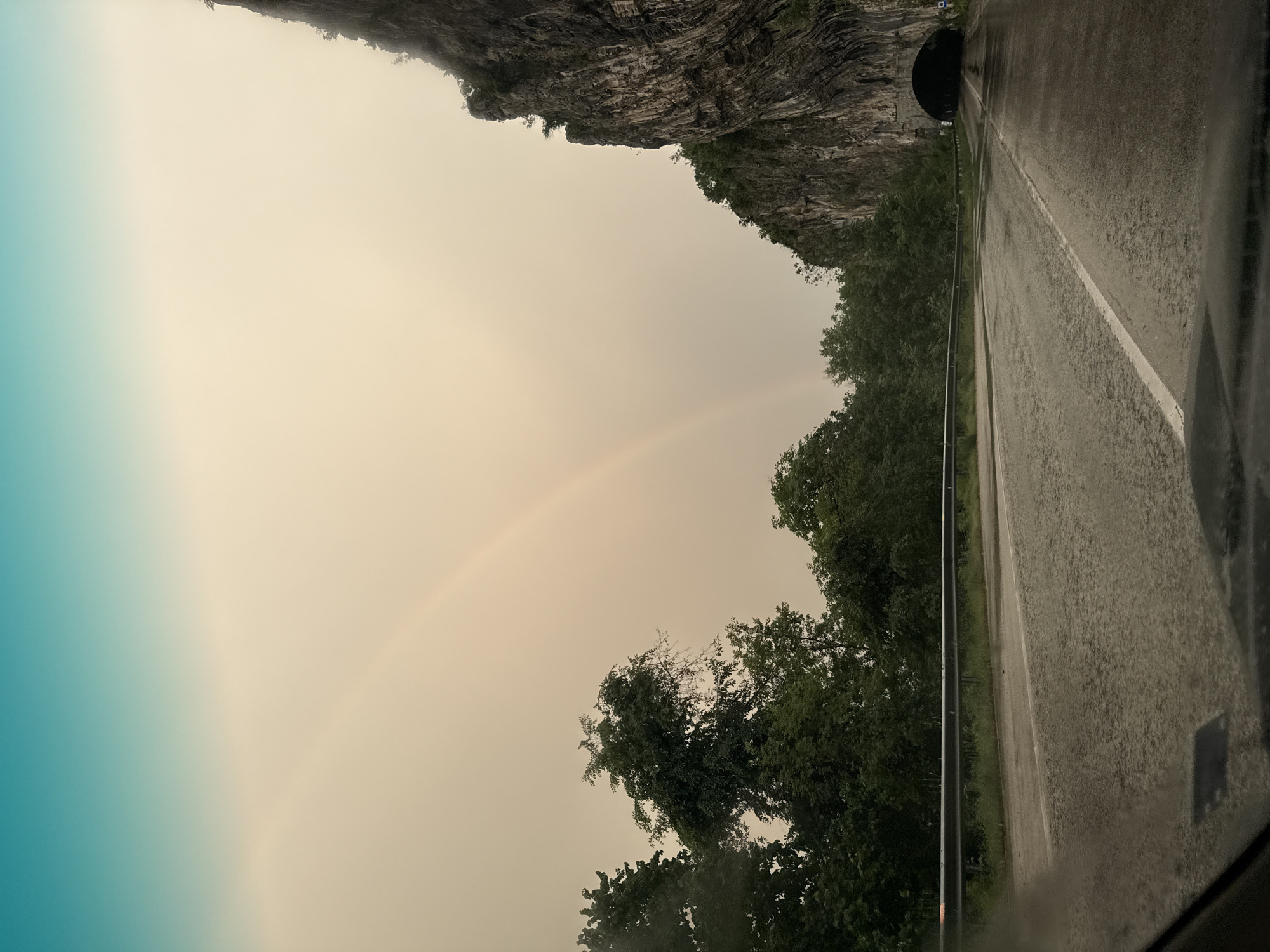 The image depicts a view from inside a vehicle driving on a road alongside a rocky cliff in Gesha, Bulgaria. The road is wet, possibly from rain. A rainbow is visible in the sky above trees alongside the road. A tunnel entrance is visible in the cliff face. The sky is overcast.