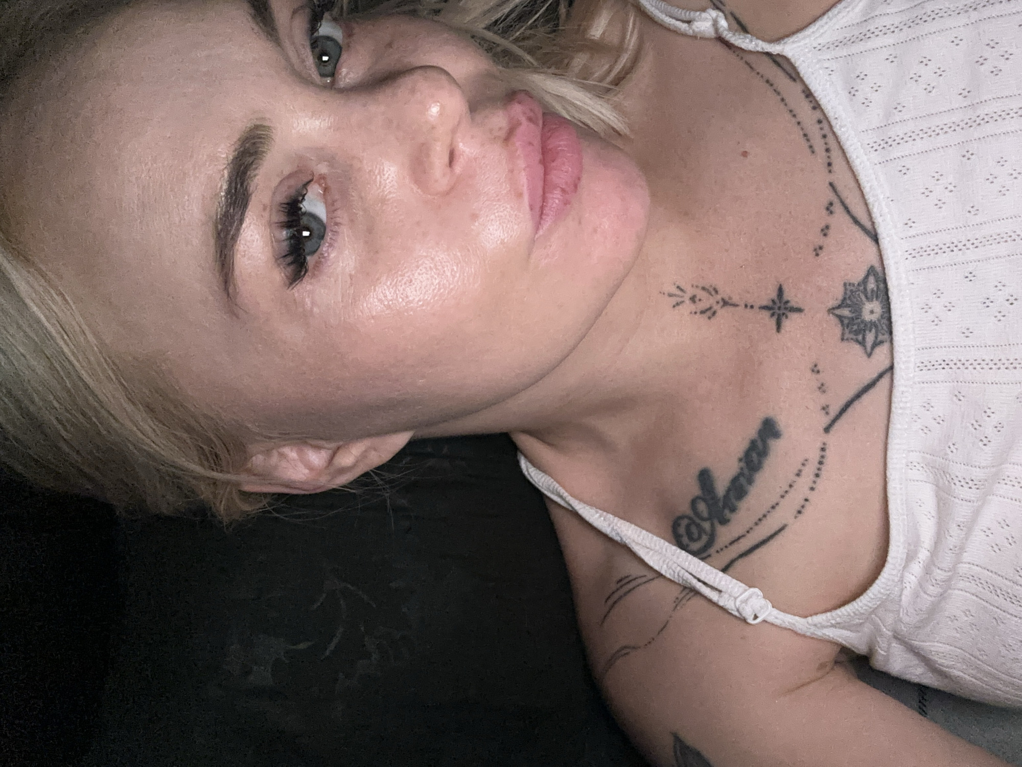 A close-up, frontal, self-taken portrait depicts an individual with light skin and blonde hair against a dark, unidentifiable background. The individual, appearing to be female, has light-colored eyes with dark, defined eyebrows and enhanced eyelashes. Their lips are full and slightly parted, showing a reddish hue. The face exhibits some texture and sheen. The individual is wearing a white, textured spaghetti-strap top with an adjustable strap visible on the left shoulder. Multiple dark-ink tattoos are present on the upper chest and right shoulder area. On the right side of the chest, a script tattoo is visible, possibly reading "Aman" or similar, accompanied by dotted lines. Centered on the upper chest, a vertical tattoo features a stylized upward-pointing element, a star-like shape, and a multi-petal design at the bottom, with additional dotted lines extending horizontally. The right ear is partially visible. The background is uniformly dark with subtle, indistinct textural variations. The specific location within the city of Unknown, Unknown, cannot be determined from the image.