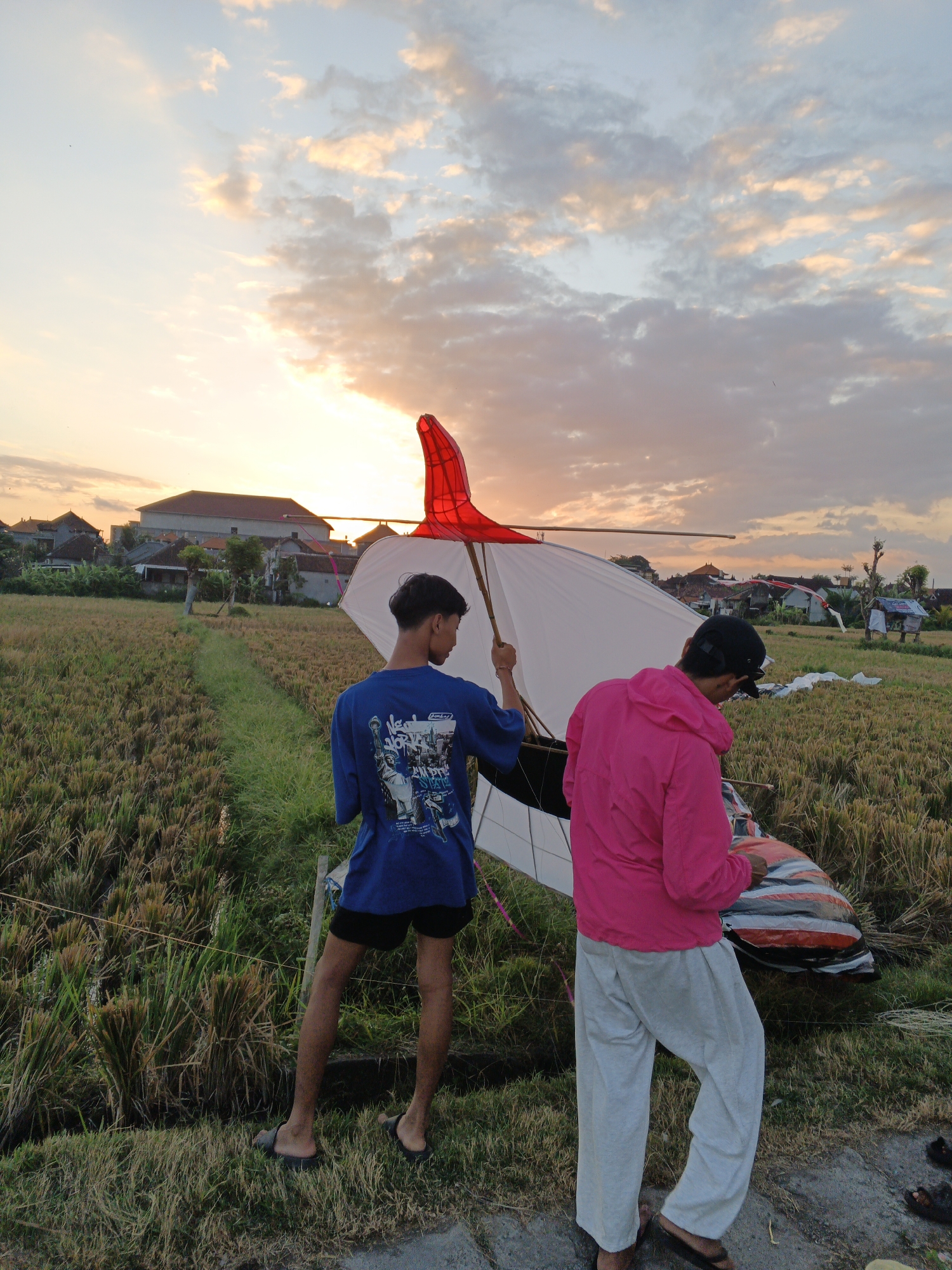 Two young men are seen in a rice field in Sukawati, Indonesia, during a sunset. The man on the left is wearing a blue t-shirt with a graphic print and black shorts. He is holding a large, white kite with red accents. The man on the right is wearing a pink hooded jacket and light gray sweatpants. He is looking down at something in his hands, possibly preparing another kite or a component of one. In the background, traditional Indonesian houses with red tile roofs are visible, nestled amongst lush greenery. The sky is painted with warm hues of orange, yellow, and pink, with scattered clouds indicating the end of the day. The rice stalks in the field are dry and golden, suggesting it is harvest season. The overall scene captures a moment of leisure and preparation for an activity in a rural Indonesian setting.
