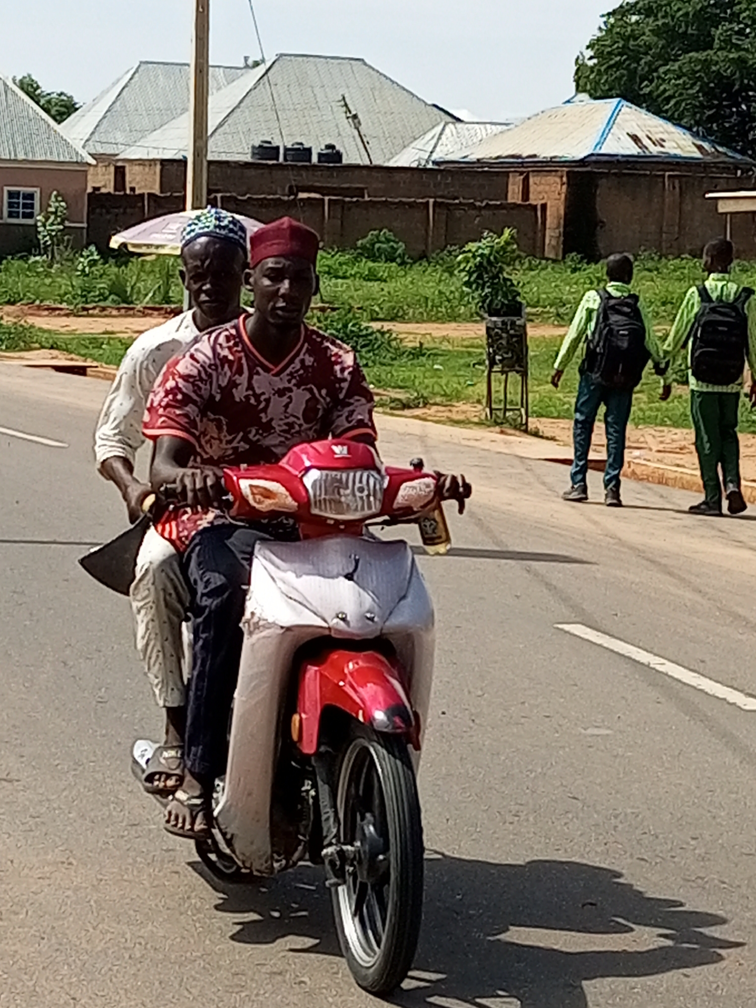 Two men are riding a red motorcycle on a paved road in Tempure, Nigeria. The man in the front is driving, wearing a red and white patterned t-shirt and a red cap. The man behind him is wearing a white long-sleeved shirt and a blue and white patterned cap. He is holding a large knife or machete with a black handle. The scene appears to be a bright, sunny day with shadows visible on the road. In the background, there are buildings with light-colored roofs, some trees, and green vegetation. Two young boys in green shirts and backpacks are walking away from the motorcycle in the background. The overall impression is a snapshot of daily life in a Nigerian town.