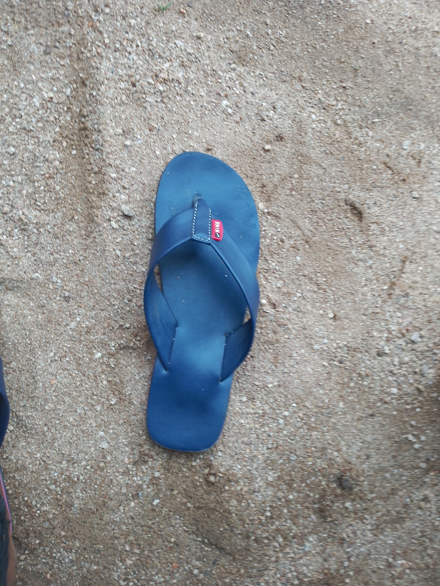 A single, blue flip-flop lies on a sandy surface. The flip-flop is oriented vertically, with the toe pointing upwards and slightly to the right. It appears to be made of a matte material, possibly rubber or faux leather. A small, red rectangular tab with white lettering, potentially a brand logo, is attached to the strap where it meets the sole. The sandy ground is composed of small grains and pebbles of various shades of brown and grey. There are some shadows and subtle variations in the texture of the sand. No people or other objects are visible in the immediate vicinity of the flip-flop. The location is not specified and cannot be determined from the image.