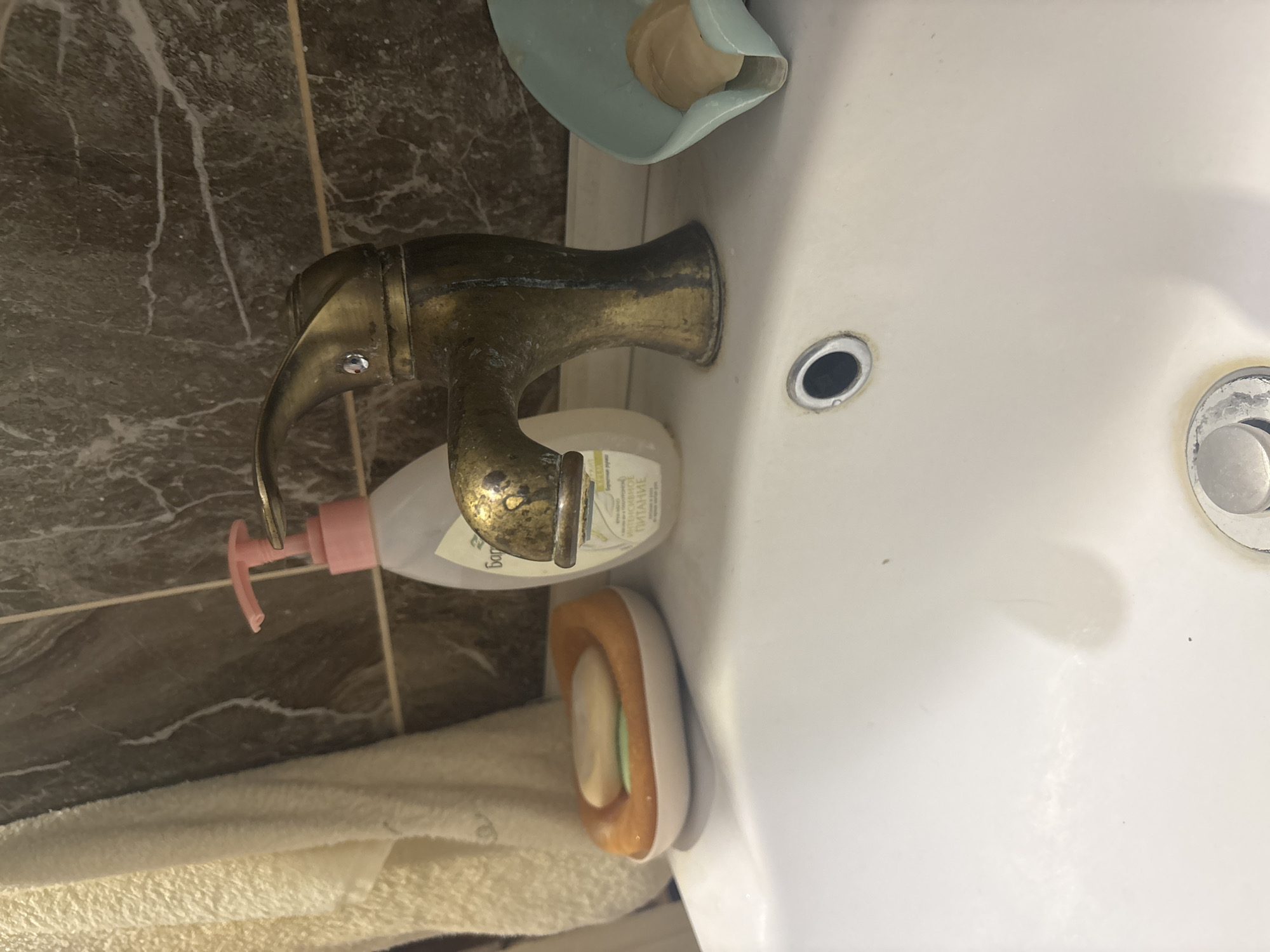 The image depicts a close-up, eye-level view of a bathroom sink area. The sink is made of white ceramic and occupies the lower portion of the frame. On the left side of the sink, there is an orange and white soap dish holding two bars of soap. Behind the soap dish, a white towel hangs vertically against a wall tiled with brown marble-patterned tiles. In the center of the image, a brass-colored faucet with an ornate design is prominent. A bottle of liquid soap with a pink dispenser is positioned behind the faucet. To the right of the faucet, a light blue soap dish contains a bar of soap. The drain of the sink is visible in the lower center of the image. The overall scene is that of a typical bathroom sink setup. The location of Березка Снт (поварово), Russia, is not discernible from the visual elements of the image.