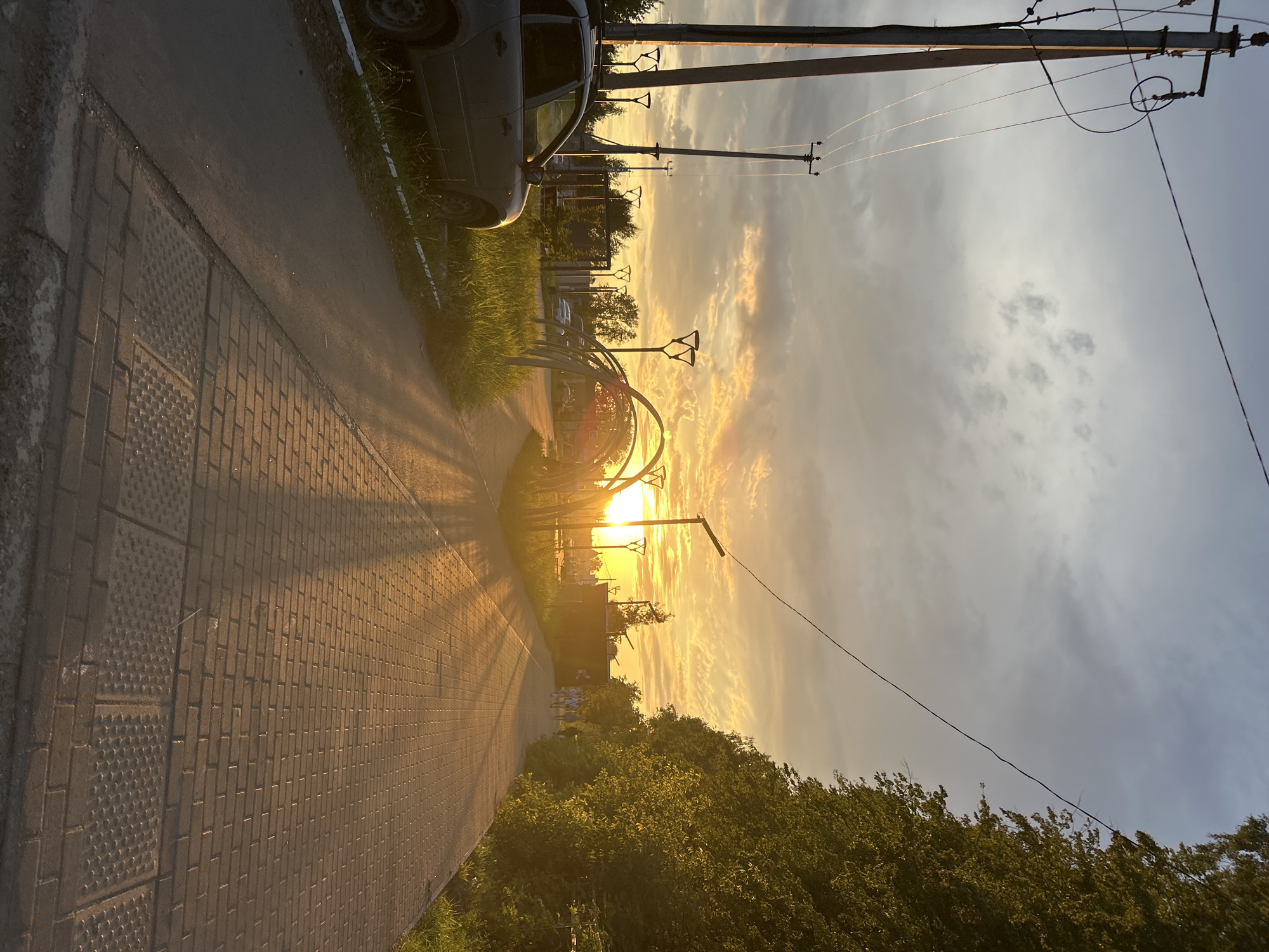 The image captures a sunset scene in Chaikovsky, Russia, along a paved pedestrian walkway. The sun is setting behind a row of trees and an arched structure, casting long shadows across the path and illuminating the sky with warm hues of orange and yellow. The walkway is made of brick pavers and runs alongside a grassy area with several modern streetlights. To the left of the walkway, a gray car is parked on a patch of asphalt adjacent to the grass. In the distance, a few people are walking along the path. Overhead, power lines stretch across the frame, supported by utility poles. The overall atmosphere is serene, with the golden light of the setting sun creating a picturesque urban landscape.