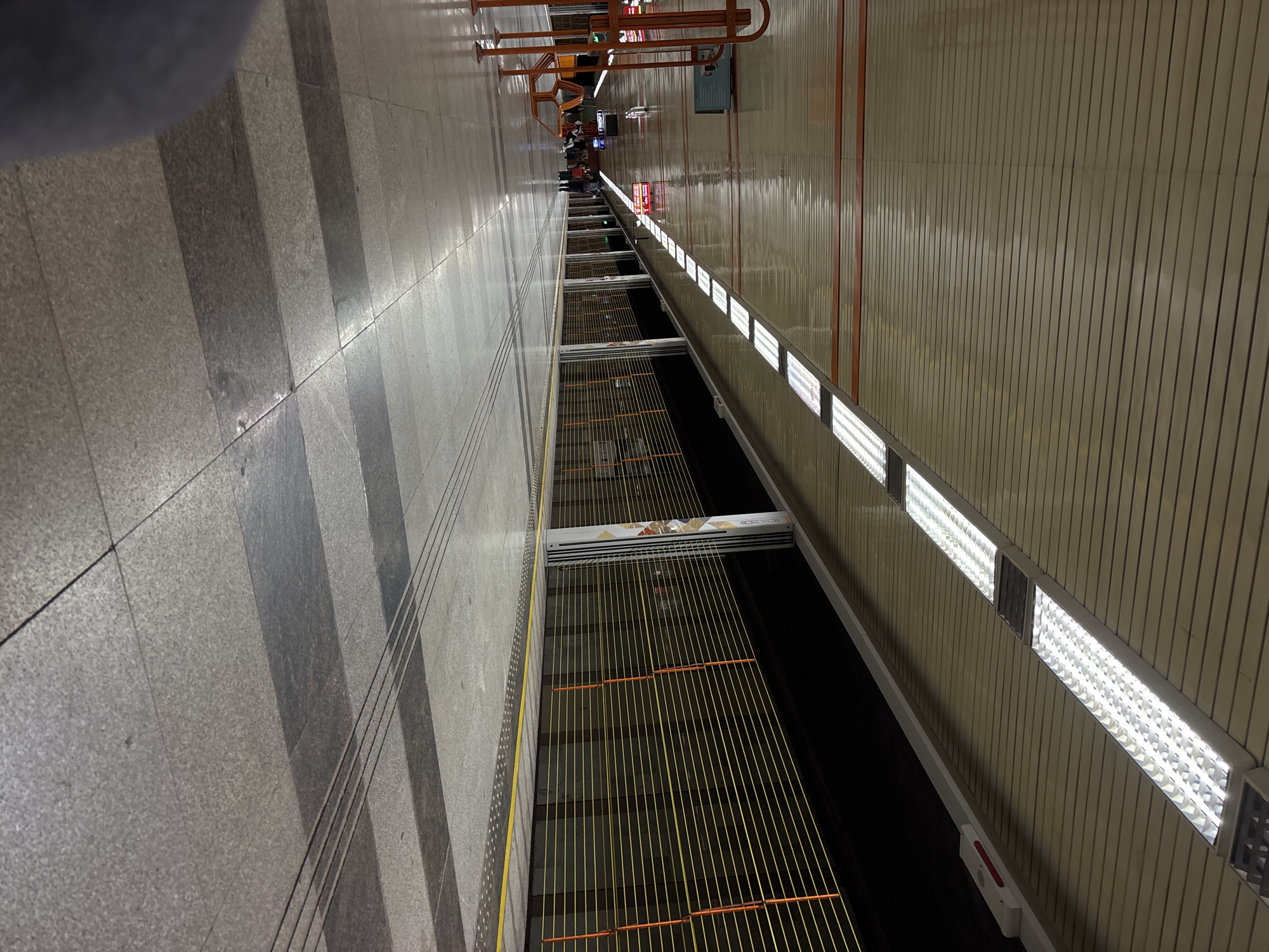 The image shows the platform of a Sofia, Bulgaria metro station. The platform is viewed from an elevated perspective, with a gray-patterned tile floor on the left and a wood-paneled wall with fluorescent lighting on the right. Between the platform edges, the train tracks run into the distance, lined with yellow safety lines. People are visible in the distance waiting on the platform. Above the train tracks there is a horizontal line of metal structures.