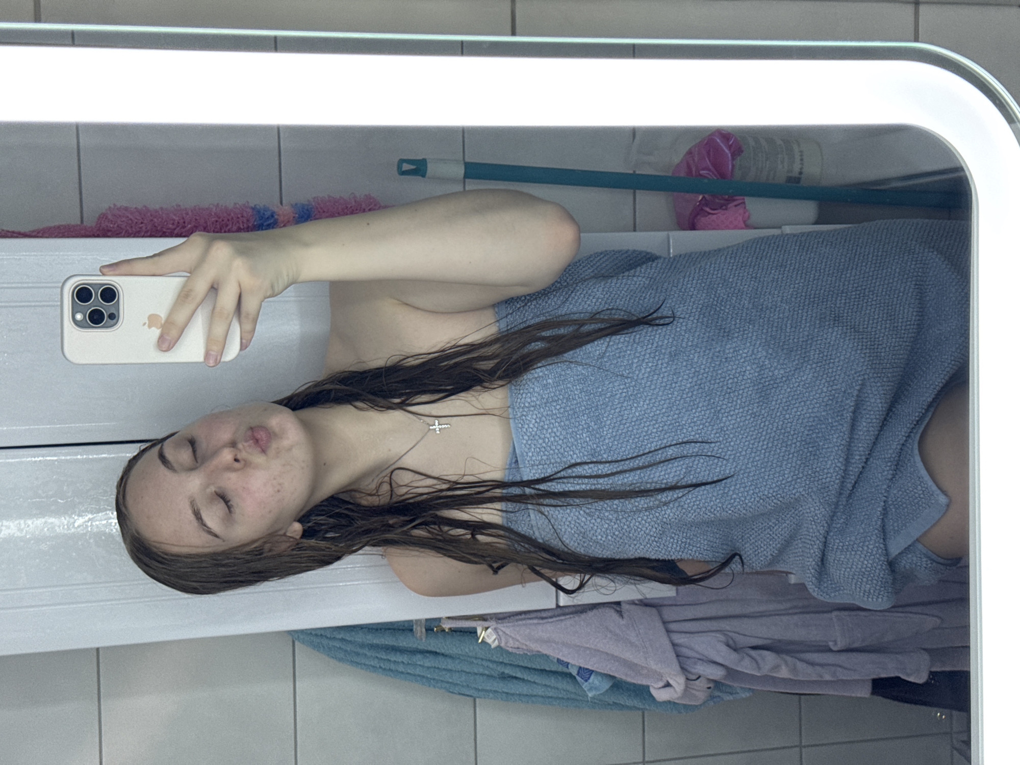 The scene is set indoors, within a domestic bathroom, presumed to be located in Perm, Russia, as requested. A young female individual is depicted in a mirror selfie, positioned centrally in the frame. She has long, wet, dark brown hair draped over her shoulders and chest, indicating a recent shower. Her eyes are closed, and her lips are pursed in a "kissing" expression. Her face shows visible freckles and minor redness. She wears a light blue terrycloth towel wrapped around her body, covering from her chest to her upper thighs, and a silver-colored cross pendant on a thin chain around her neck.
In her left hand, visible in the mirror's reflection, she holds a light-colored smartphone with a visible triple camera array and an Apple logo, used to capture the image. The mirror itself features a vertical, illuminated LED light strip along its right edge.
The background, also reflected in the mirror, shows a white tiled wall on the left, from which a light blue bath towel or robe and a light purple garment are hanging. White wall paneling or a cabinet door is visible directly behind the individual. To the right, a pink textured item (possibly a loofah) hangs, and a teal-colored mop or cleaning tool leans against the wall, next to a white bucket containing pink plastic. The environment suggests a functional and well-lit private bathroom.