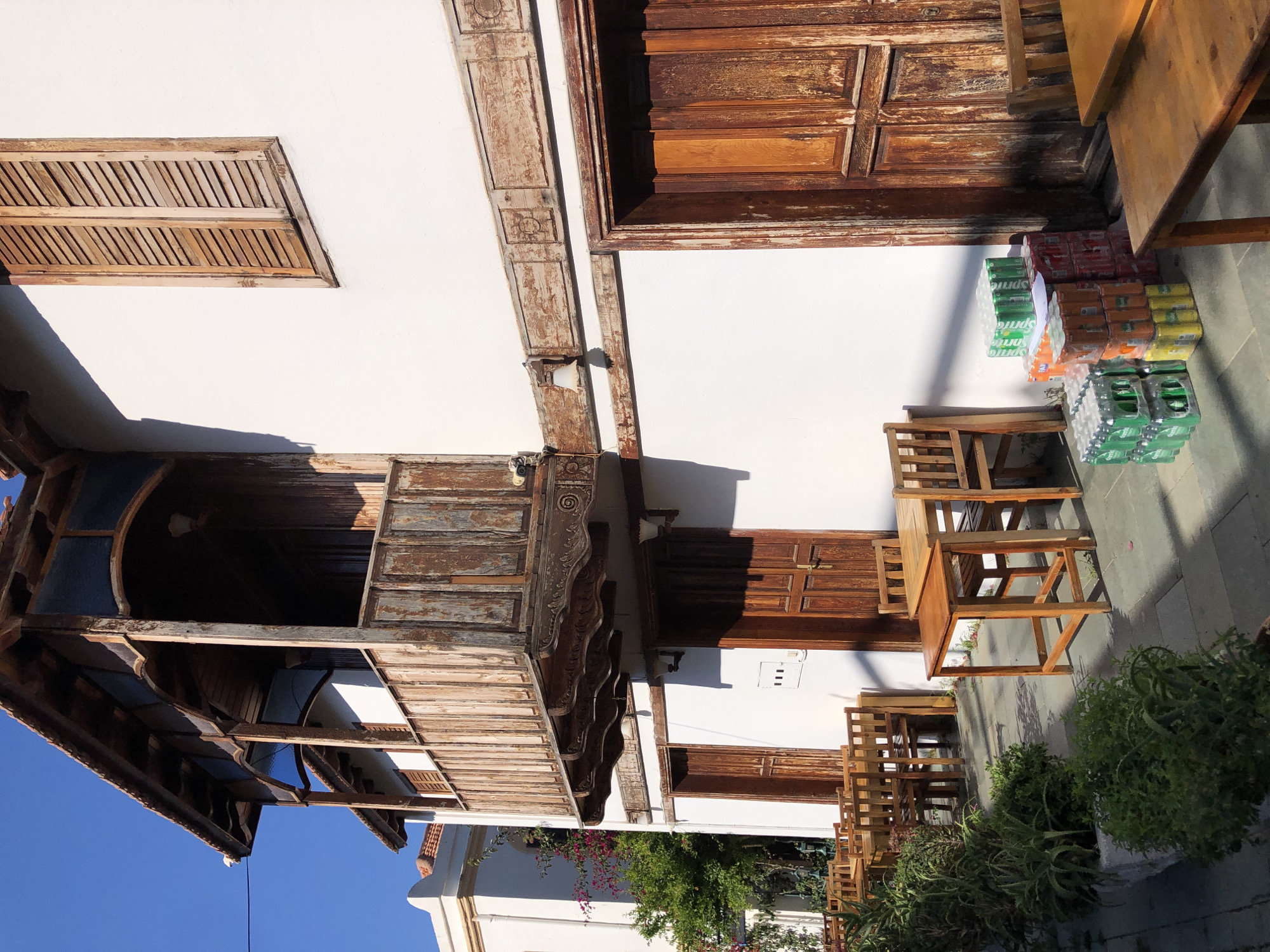 The image depicts a street scene in Kaş, Turkey, focusing on the exterior of a traditional building with weathered wooden architectural details and a white facade. A covered balcony with intricate wooden carvings and vertical planks is a prominent feature, showing signs of age and peeling paint. Below the balcony, two dark wooden doors are visible on the white wall, along with an electrical box and two outdoor light fixtures.
To the left of the building, a row of empty wooden tables and chairs is set up on a paved walkway, suggesting an outdoor seating area for a cafe or restaurant. Lush green plants and bushes are arranged in planters along the walkway, adding a touch of nature to the scene. In the foreground, a stack of beverage crates, including green Sprite and orange Fanta cans, is placed on the ground next to a wooden table and chairs. The sunlight casts strong shadows, indicating a bright, sunny day. In the background, another building with a tiled roof and wooden shutters is partially visible, hinting at the architectural style of the town. The overall impression is one of a charming, historic street in a Mediterranean coastal town.