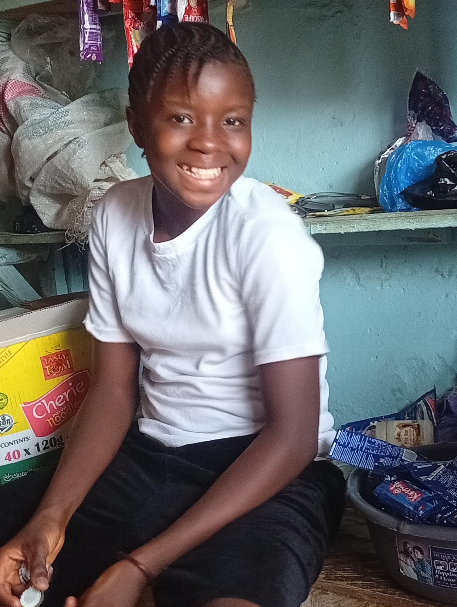 A young dark-skinned person with braided hair sits indoors wearing a white t-shirt and dark pants. They are smiling at the camera and hold a bottle cap in their left hand. A yellow box labeled "Tasty Tom Cherie Instant Noodles" is visible to the left. The background includes a light blue wall, a shelf with various items, and a container filled with packaged goods on the right. The location appears to be a small shop or storage area, possibly in a city, but the specific city and country are unknown.