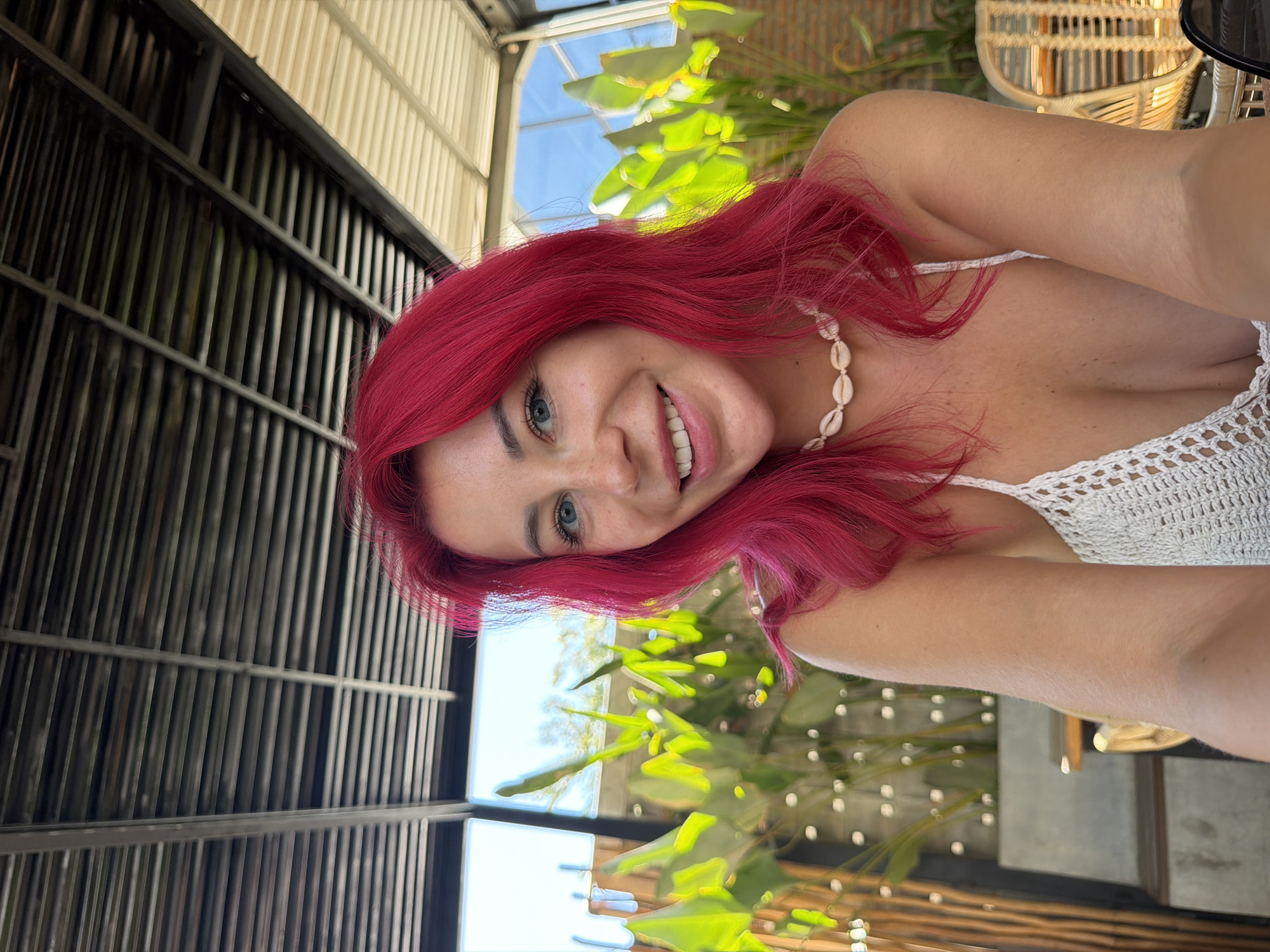 This media file features a young woman with vibrant pink hair. She is wearing a white crocheted top and a seashell necklace. The woman is smiling and looking towards the camera, with a relaxed and joyful expression. Her blue eyes are clearly visible, and she has a smattering of freckles across her nose and cheeks.
The setting appears to be an outdoor or semi-outdoor space, possibly a cafe or restaurant, given the presence of chairs made of woven material and lush green foliage. The background includes a textured ceiling structure, likely part of the building's roof, and metal grating on the left side, suggesting an open-air design. Sunlight filters through the plants and the structure, indicating it's daytime. The overall ambiance is casual and tropical, aligning with the provided location context of Kuta, Indonesia. There is no visible text in the image. The woman's pose and the selfie-style framing suggest she is taking the photo herself.