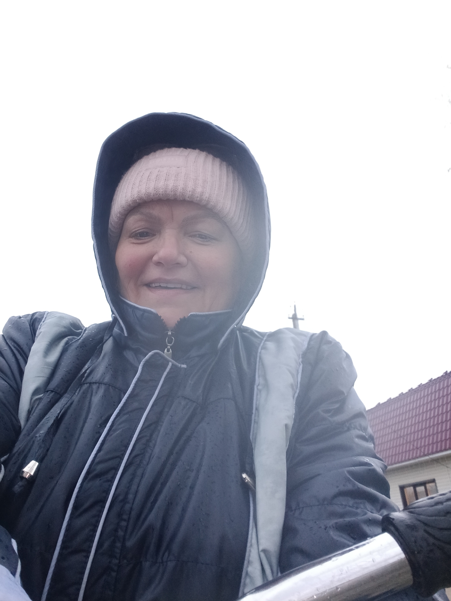 A selfie-style portrait captures a smiling, middle-aged woman outdoors in what appears to be Красный Маяк, Russia. She is dressed warmly for inclement weather, wearing a dark-colored, possibly navy or black, hooded jacket with light grey piping and shoulder panels. Numerous small water droplets are visible on her jacket, indicating it is raining or has recently rained. Underneath the hood, she wears a light pink or beige knitted beanie hat. Her expression is cheerful and content as she smiles directly at the camera.
The photo is taken from a low angle, looking upwards towards her face and the sky. The background is predominantly a bright, overcast sky, suggesting daytime. To the right of the woman, a portion of a building with a distinct reddish-brown tiled roof is visible, suggesting a residential or urban setting. A slender, cross-shaped structure, possibly a utility pole or antenna, can be faintly seen in the mid-ground. In the bottom right corner, a reflective metal bar, resembling a bicycle handlebar or a railing, is present. The woman appears to be engaged in an outdoor activity, possibly cycling given the presence of the bar. No visible text is present in the image.