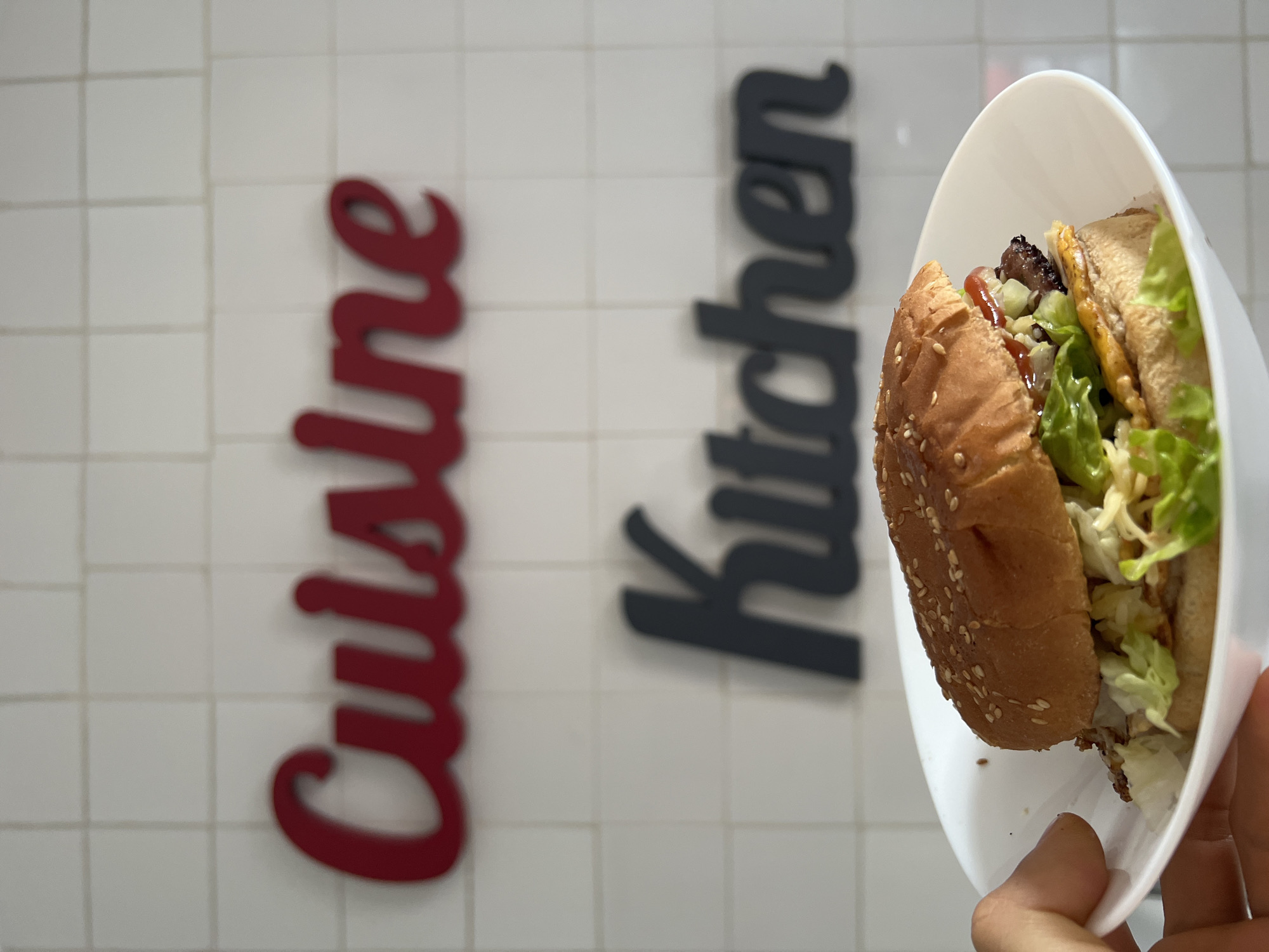 The image shows a hamburger in a white paper container, held in someone's hand. The hamburger contains a sesame seed bun, lettuce, a patty, and sauce. Behind the burger, a white tiled wall is visible with the words "Cuisine" in red cursive and "Kitchen" in dark gray cursive. The image was likely taken indoors in a restaurant or kitchen. While the image provides visual context and branding cues, it does not provide any direct information regarding the city of Madrid, Spain. Therefore, I cannot definitively state this image was taken in Madrid, Spain.