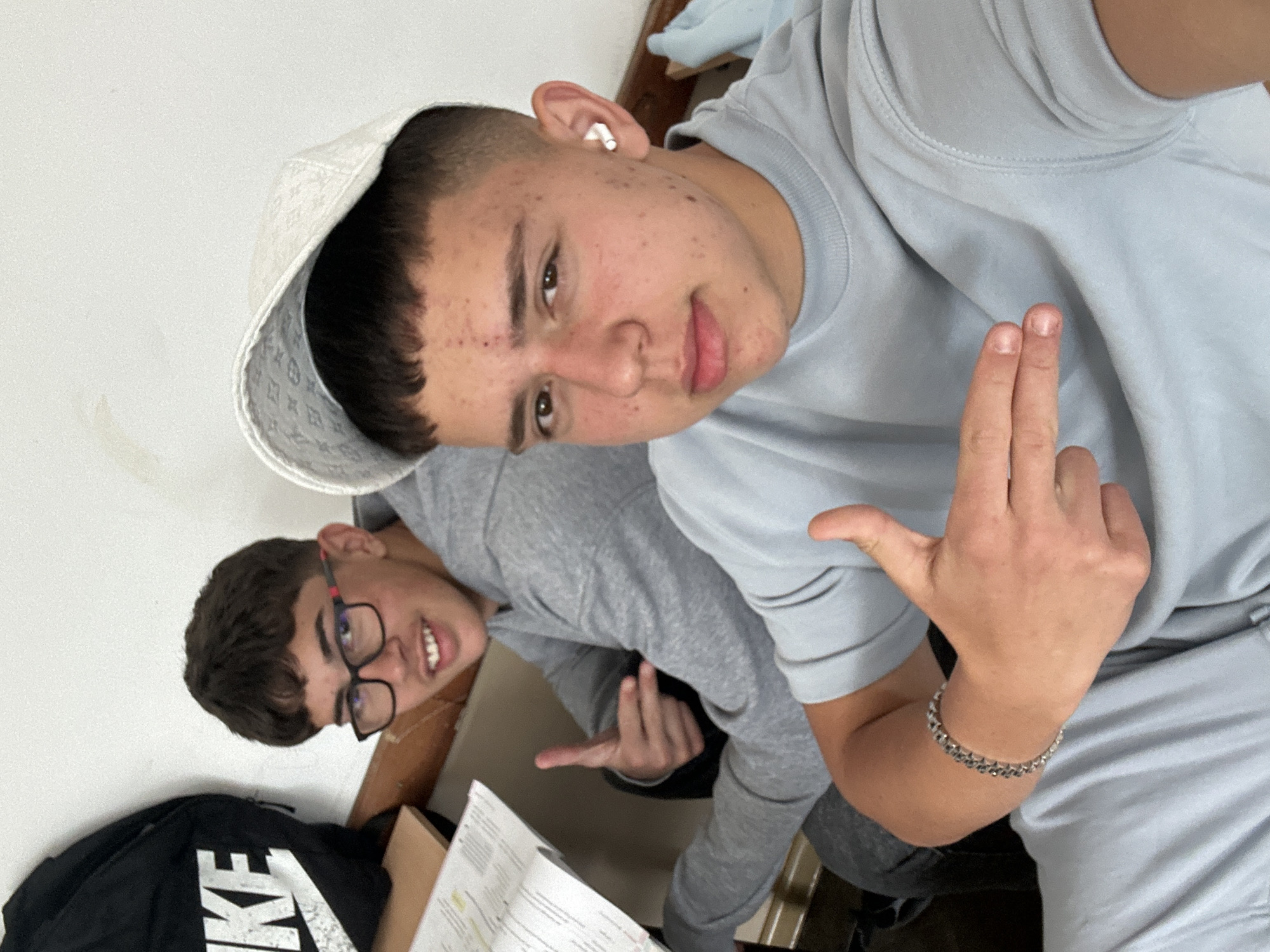 Two males are pictured indoors. The male in the foreground, wearing a white hat and light blue shirt, makes a hand gesture. A silver bracelet adorns his wrist. The second male, wearing glasses and a gray shirt, is positioned behind him and also makes a hand gesture. A black Nike bag and papers are visible. Location is indeterminate and there is no evidence it's in Zlatograd, Bulgaria.