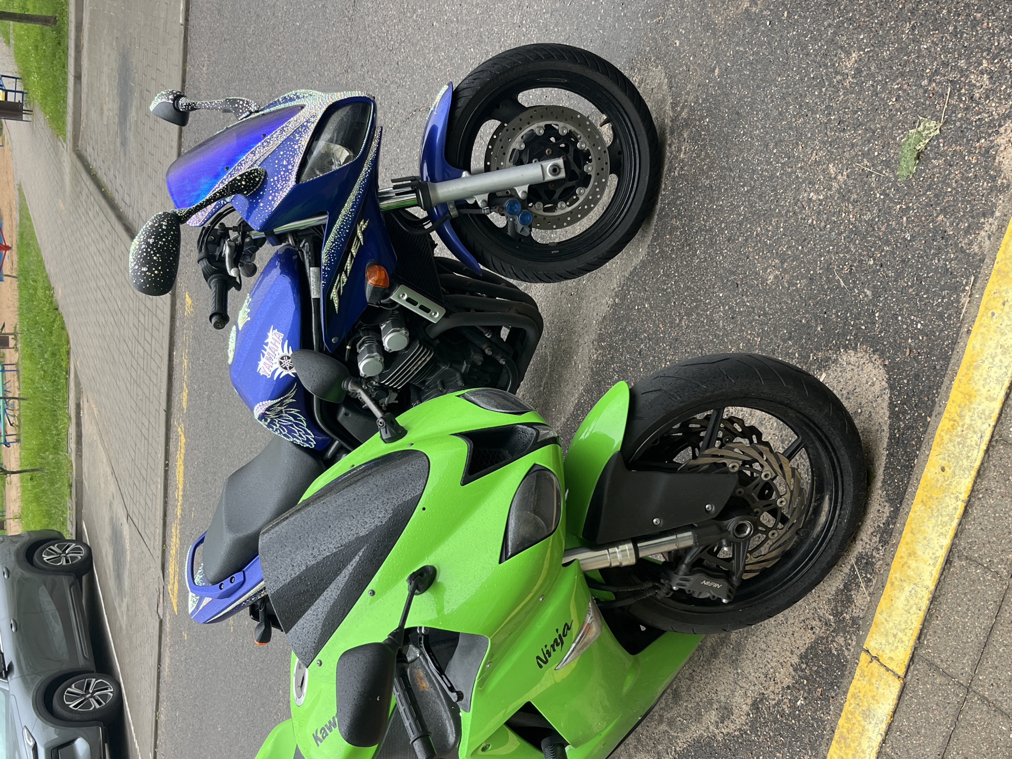 This is a medium shot, eye-level, outdoor photograph of two parked motorcycles on an asphalt surface with a yellow painted curb in the foreground and a paved walkway behind. The motorcycle on the left is bright green and has the word "Ninja" written on its side. The motorcycle on the right is blue and appears to be decorated with glitter or small reflective stickers. Both motorcycles have wet surfaces, suggesting recent rain. In the background, a grey car is parked, and beyond that, a small playground area with a red slide and blue and yellow equipment is visible. The overall environment appears to be a residential area or public parking lot. No people are visible in the image. The image was taken in Minsk, Belarus.