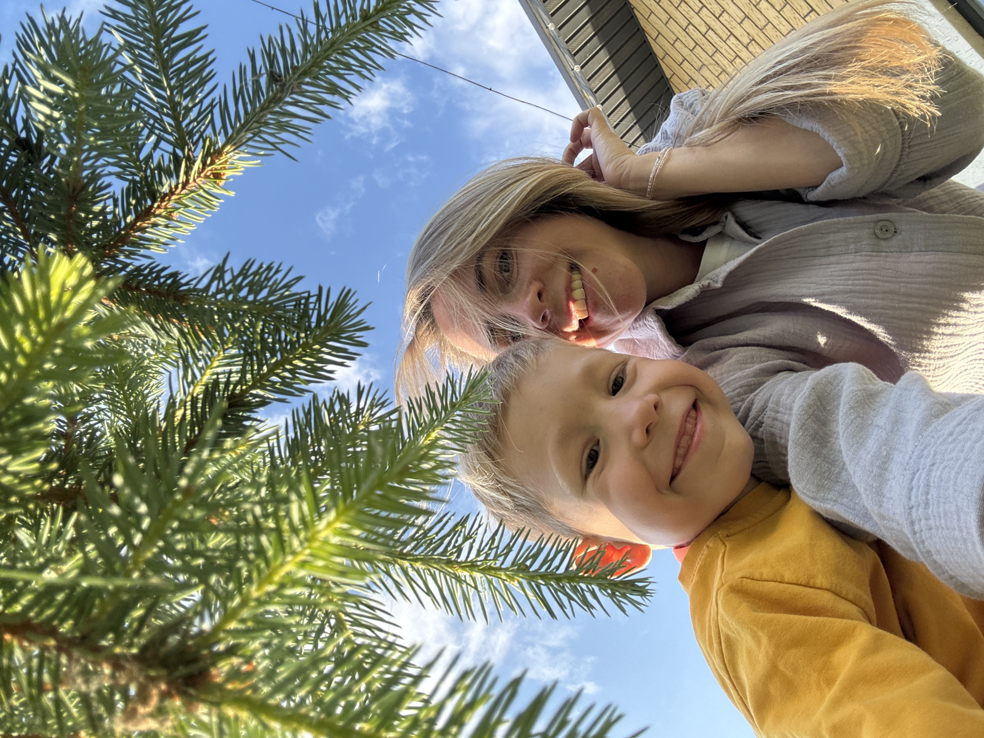 In an outdoor, low-angle, selfie-style photograph, a young boy with fair hair smiles widely, wearing a yellow long-sleeved shirt. Next to him, a woman with blonde hair and fair skin smiles, her head tilted and her hand touching her hair. Both are positioned in the foreground. Overhead, the branches and needles of a coniferous tree create a frame, with a bright blue sky and scattered white clouds visible through the foliage. In the background, the side of a building with beige brickwork and a dark-grey roof is visible. The scene appears to be in Chaikovsky, Russia, under natural sunlight.