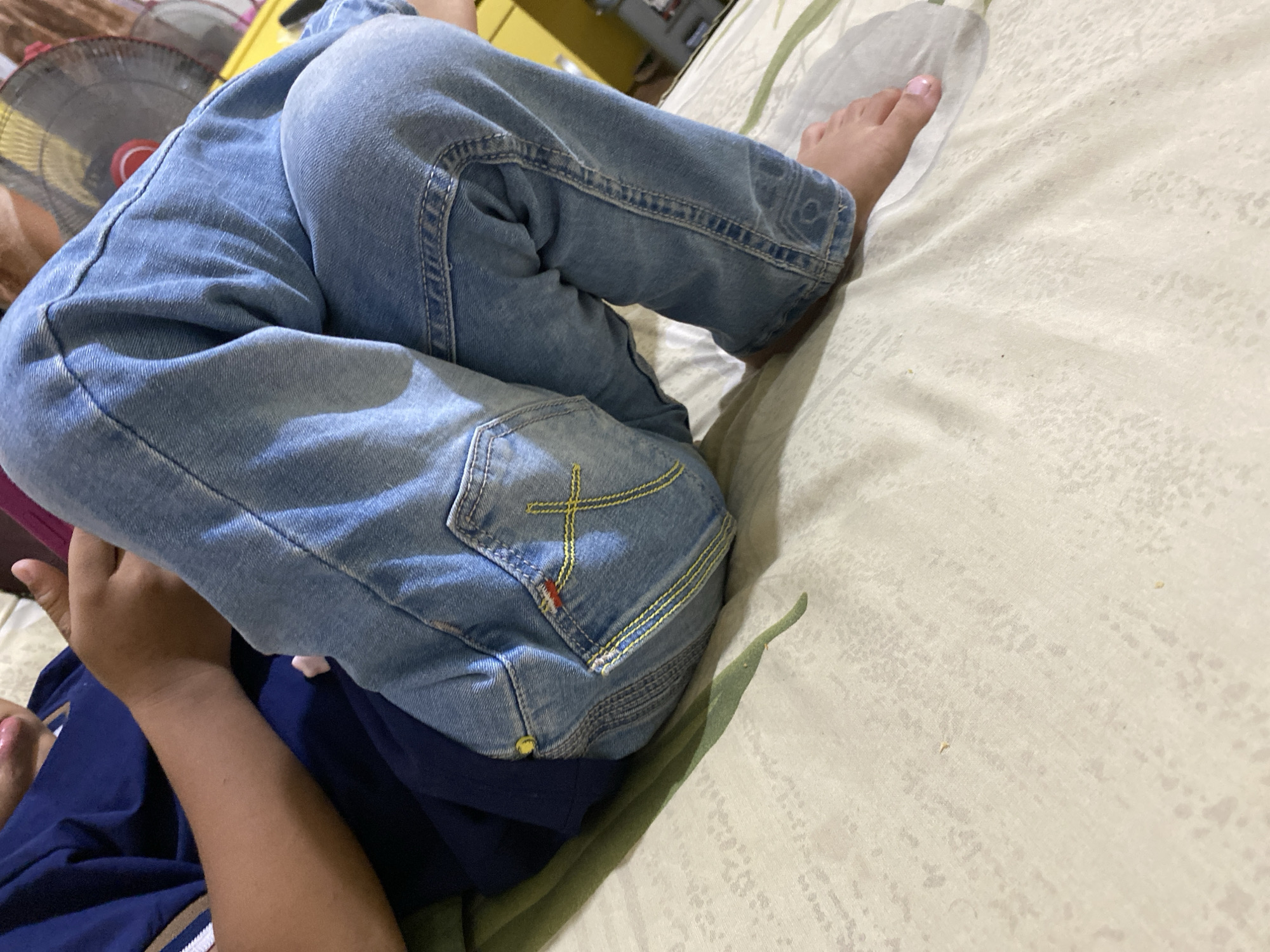 The image shows a close-up of a person, likely a child, wearing a dark blue t-shirt and light blue jeans, sitting on a bed. The jeans have decorative yellow stitching on the back pocket. The person's hands are visible, holding a pink object, possibly a phone or tablet. The background is blurred, but a yellow object and part of a fan are discernible. The environment appears to be an indoor setting, possibly a bedroom. The lighting is moderate, creating some shadows. There are no specific city or location markers visible in the image.