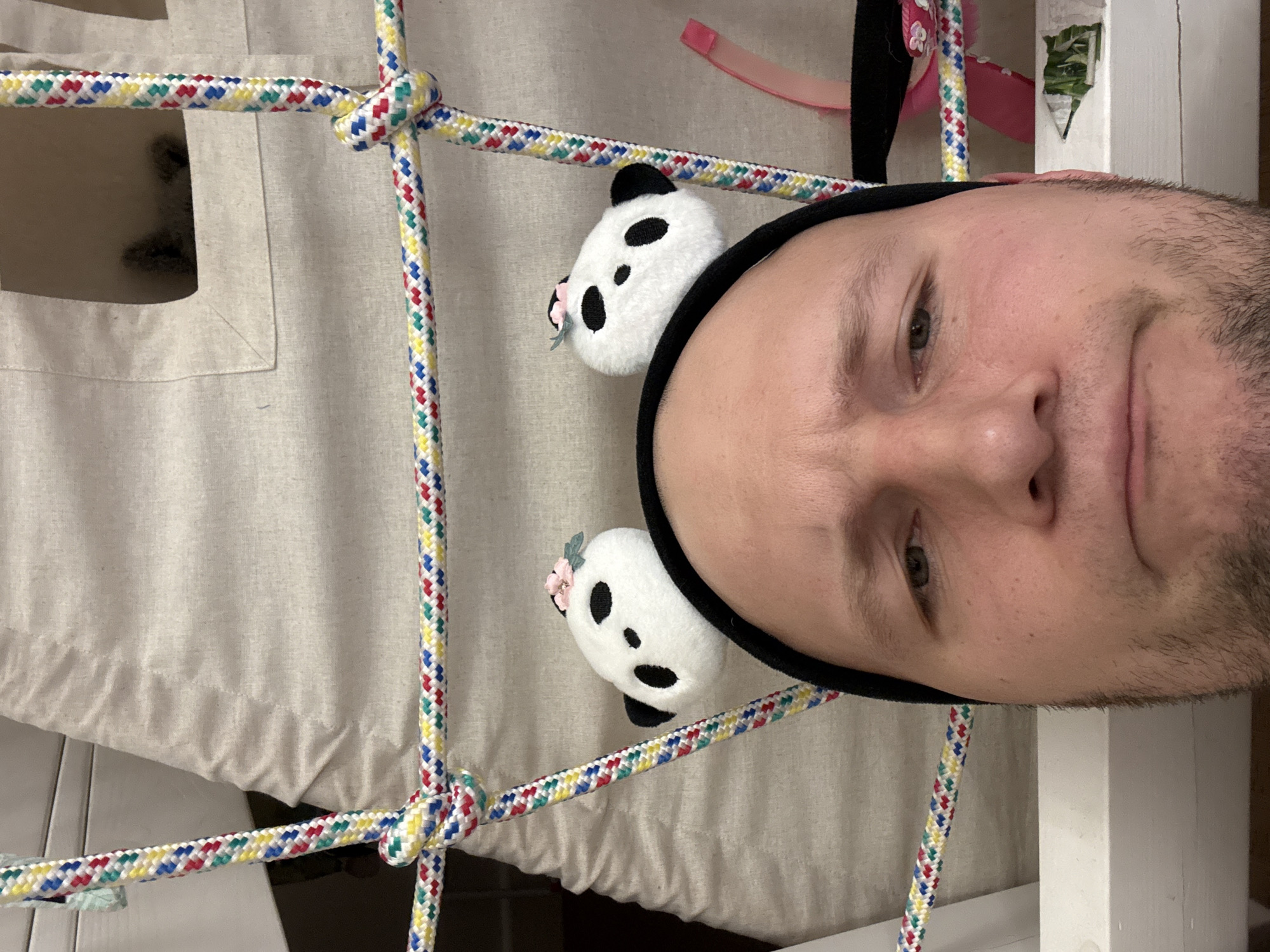 A person with a black headband is positioned to the right of a rope net with two panda-shaped decorations. The net is set against a light-colored fabric backdrop with a visible opening. Additional decorative elements are subtly present in the background, including a pink ribbon and a green-patterned sticker. The setting and location of this image are not discernible based on the provided visual information.