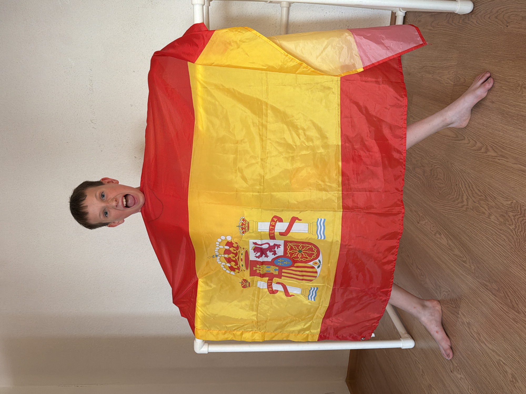 In the foreground of the image, a young boy is peeking out from under a large Spanish flag. The flag, predominantly red and yellow with the Spanish coat of arms displayed prominently, is draped over a white structure that appears to be a rack or a small bed frame. The boy has short brown hair and is wearing a blue t-shirt. His eyes are wide open and he has a joyful, excited expression on his face, with his mouth open in a shout or laugh.
His body is largely obscured by the flag, but one of his feet is visible to the right, resting on a wooden floor. The foot shows some red marks. The background consists of a textured, light-colored wall and the wooden floor. The scene appears to be indoors, in what could be a bedroom or a play area. The lighting suggests it is daytime. The overall impression is one of playful energy and enthusiasm, possibly related to a sporting event or a national celebration. There is no visible text in the image, other than the text on the Spanish coat of arms on the flag. The location context is Yekaterinburg, Russia.