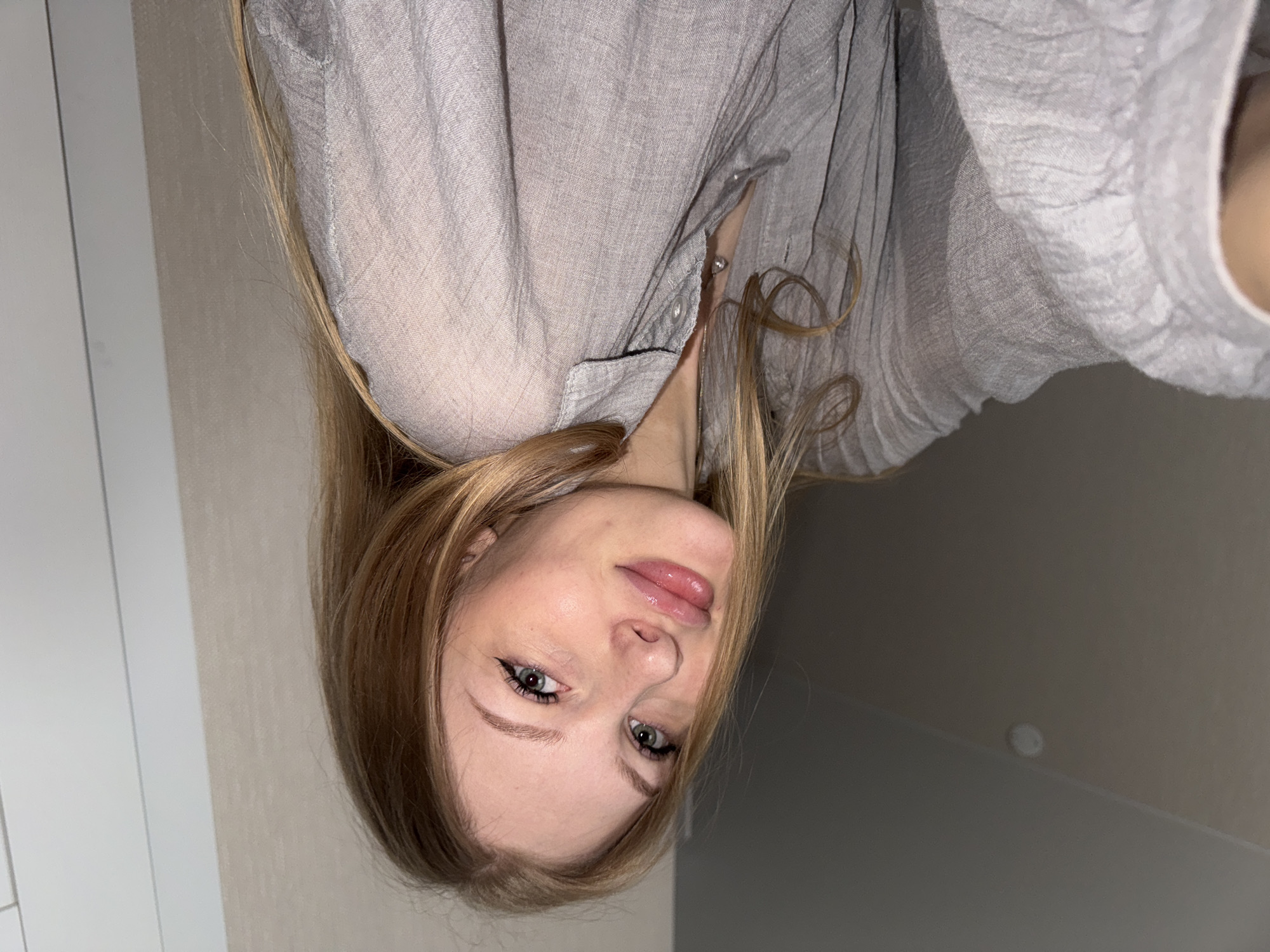A woman with blonde hair and light-colored eyes is captured in a close-up selfie. She is wearing a light grey, loose-fitting shirt with ruffled fabric. Her lips are full and glossy, and she has subtle makeup on her eyes, including eyeliner. The background appears to be an indoor setting with a plain, textured wall and a white vertical element, possibly a door frame. The lighting is soft, creating a diffused illumination. There is no identifiable information suggesting the image was taken in Perm, Russia.
