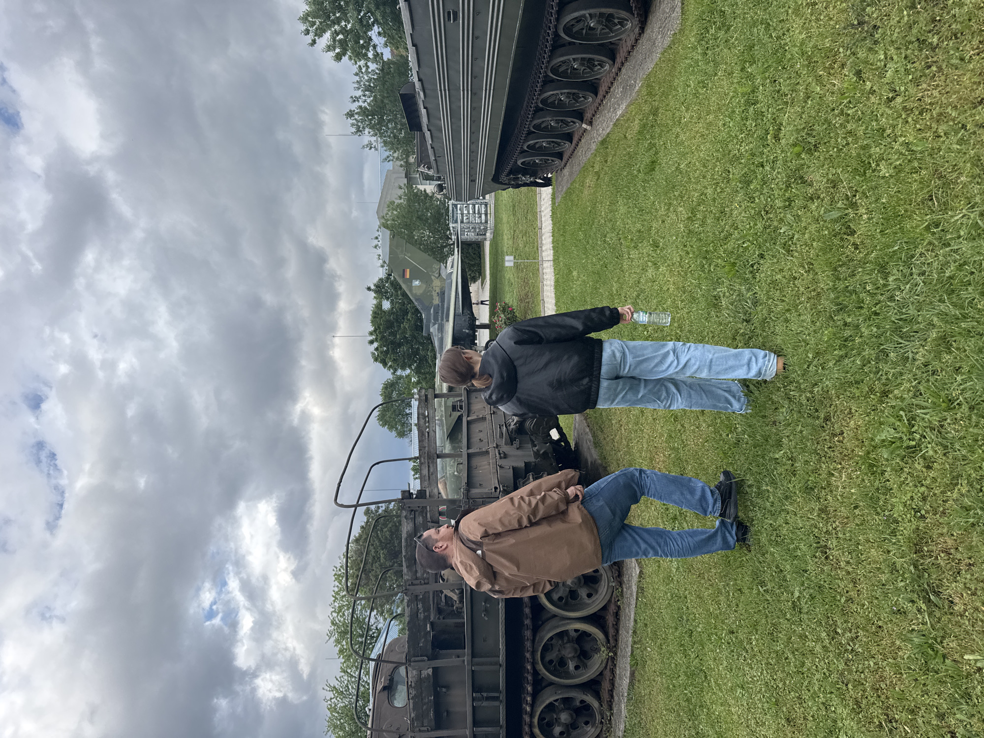 The image shows two people, likely male, standing next to military vehicles in an outdoor environment. One person is wearing a brown jacket, jeans, and black shoes. The other is wearing a black jacket, jeans, and holding a plastic water bottle. The vehicles are on display on a patch of green grass under a cloudy sky. While I can't definitively confirm the location as Sofia, Bulgaria, the presence of military vehicles and the European architectural style of buildings visible in the background is consistent with that region.