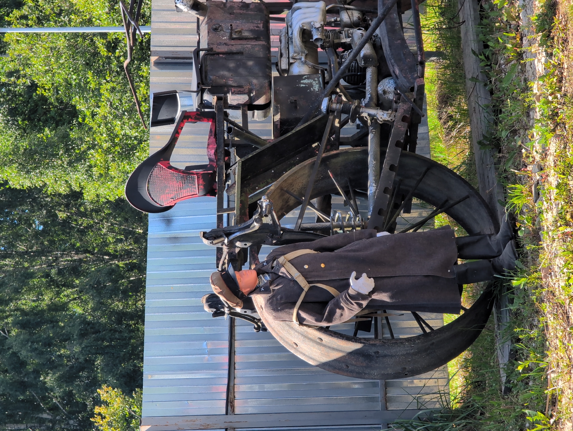An outdoor scene in Novoe Devyatkino, Russia, features a mannequin dressed in a historical military-style uniform, standing next to a large, custom-built vehicle that appears to be a stationary display. The mannequin wears a brown trench coat, a cap, white gloves, and black boots. It is positioned to the left of the vehicle. The vehicle is dark-colored and appears to be made of metal, with a large, weathered wheel on the left side. It has a seat with a red mesh backrest and a black cushion, as well as visible engine components. A corrugated metal fence serves as a backdrop, with green trees and foliage visible beyond it. The ground is covered in grass and gravel. The lighting suggests it is daytime with sunlight casting shadows.