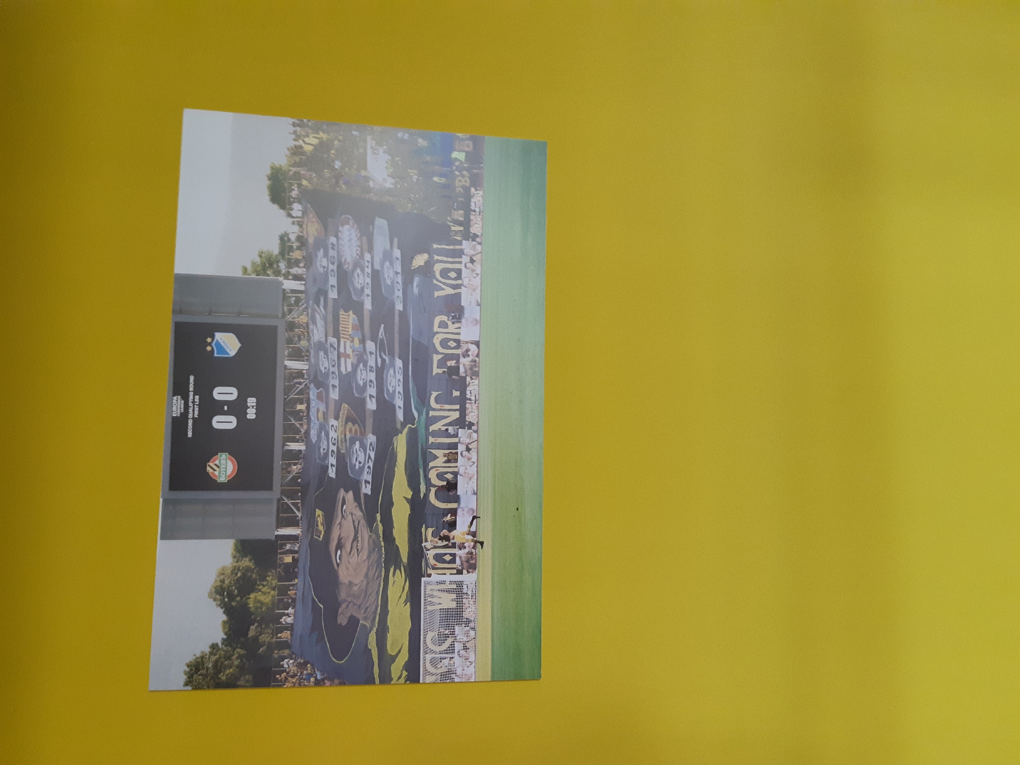 The image shows a photograph of a soccer stadium, most likely the Botev 1912 Football Stadium located in Plovdiv, Bulgaria. The scoreboard shows the game tied at 0-0 with a time of 00:19 (likely the minute of the game). A large banner spans the length of the stadium, it has a color scheme of yellow and dark blue. The banner includes the inscription "WE'RE COMING FOR YOU" and a series of years related to significant club achievements or founding dates (1962, 1967, 1968, 1972, 1984, and 2013).