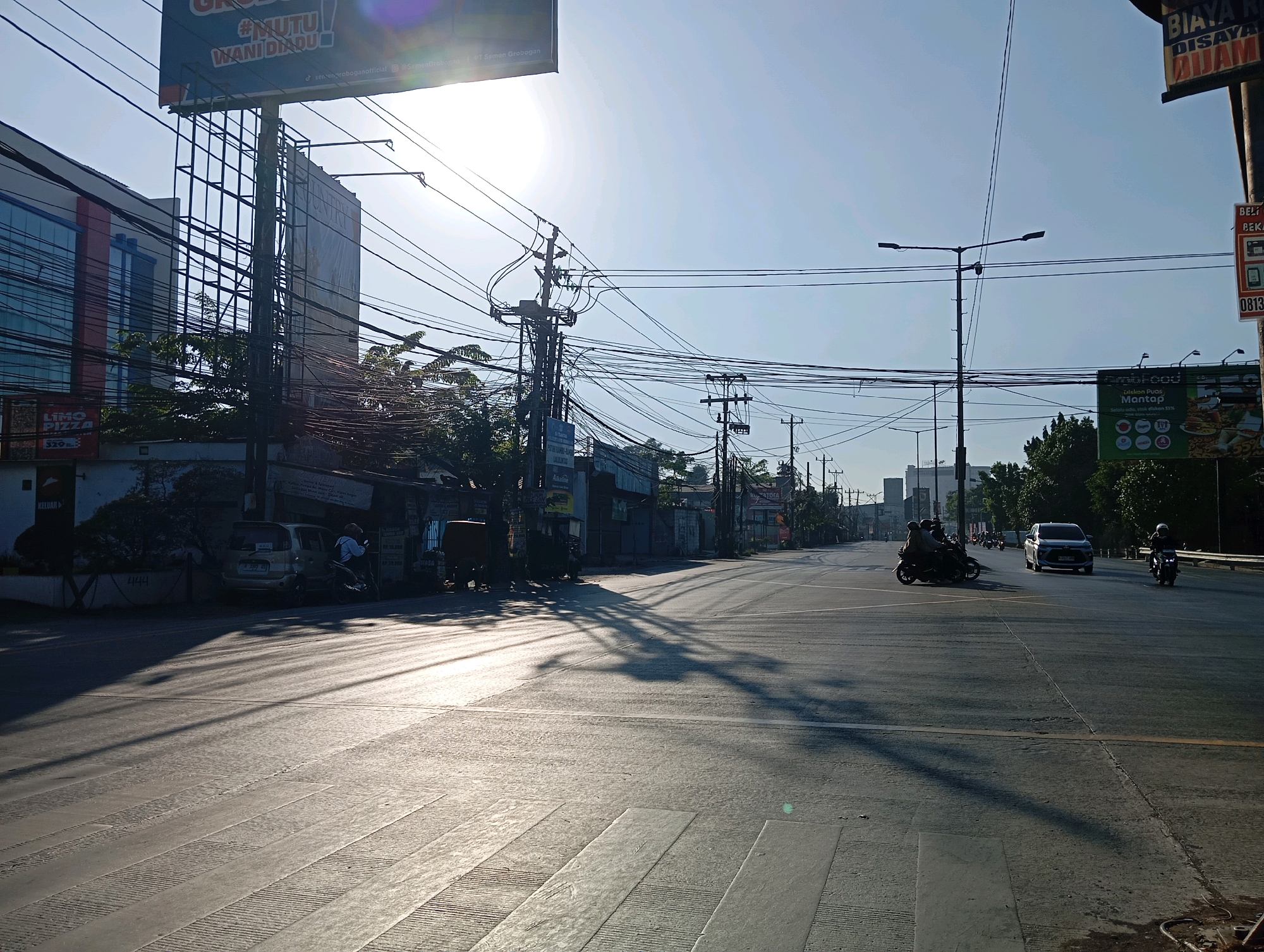 The image shows a street scene in the city of Semarang, Indonesia, likely in the early morning or late afternoon, given the long shadows cast across the road. The sun is bright and low in the sky, contributing to the strong contrast and lens flare visible in the upper portion of the image. The street is paved and appears to be a multi-lane road with visible lane markings and a pedestrian crossing.
On the left side of the road, there is a building with a modern facade featuring blue-tinted windows and a red and white stripe design. A tall billboard structure stands in front of this building, with a partial advertisement visible at the top. Below the billboard, a sign for "LIMO PIZZA" with a price of "5.29" is also visible. Several utility poles with a dense network of electrical and telecommunications wires are prominent features along the roadside, creating a visually complex aerial structure.
Further down the street, on the left, a few parked cars and motorcycles are present. One car, a silver minivan, is visible with people on a motorcycle behind it. To the right, more buildings and shops line the street, some with awnings and signs, though many are not clearly legible.
The road itself has a few vehicles on it. In the middle distance, there are several motorcycles with riders, and a silver car is traveling in the same direction. A single motorcycle with a rider is also moving in the opposite direction on the right side of the road. The traffic appears light.
The overall environment is urban, with a mixture of modern and older structures. The presence of numerous power lines is a notable characteristic of the scene. The atmosphere appears calm, with no overt signs of activity or congestion.