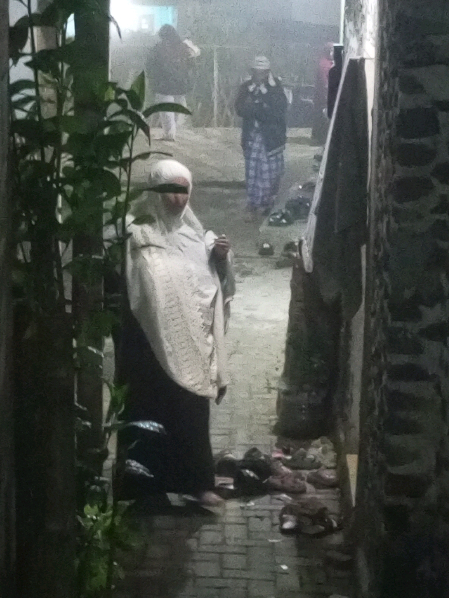An out-of-focus, nighttime image depicts an alleyway in Andongsili, Indonesia. In the foreground, a person wearing a white hijab and dark clothing stands to the left, partially obscured by greenery. Further down the alley, another individual in a checkered sarong and a dark jacket with a hat is visible. The ground is paved with bricks and is littered with several pairs of sandals. A hazy, misty atmosphere pervades the scene, suggesting damp or foggy conditions. The alley walls are constructed of stone.