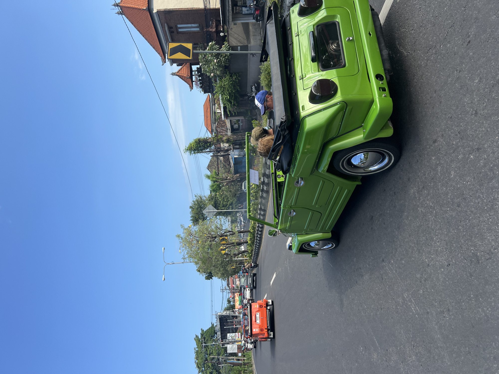 The image shows an eye-level view from the side of a bright green open-top vehicle moving along a busy street in Denpasar, Indonesia. The vehicle appears to be a Volkswagen Type 181, colloquially known as a "Thing." Two people occupy the car: the driver, wearing a blue and white baseball cap, and a passenger with a brown hat. The road surface is asphalt and appears to be in good condition. Other vehicles, including an orange Jeep, are visible in the traffic ahead. The surrounding environment features buildings of varying heights and architectural styles, palm trees, and street lights. A yellow road sign is visible near a building. The sky is clear and blue, suggesting a sunny day. Overall, the scene captures a slice of daily life in a bustling urban setting, likely in a tourist area due to the type of car.