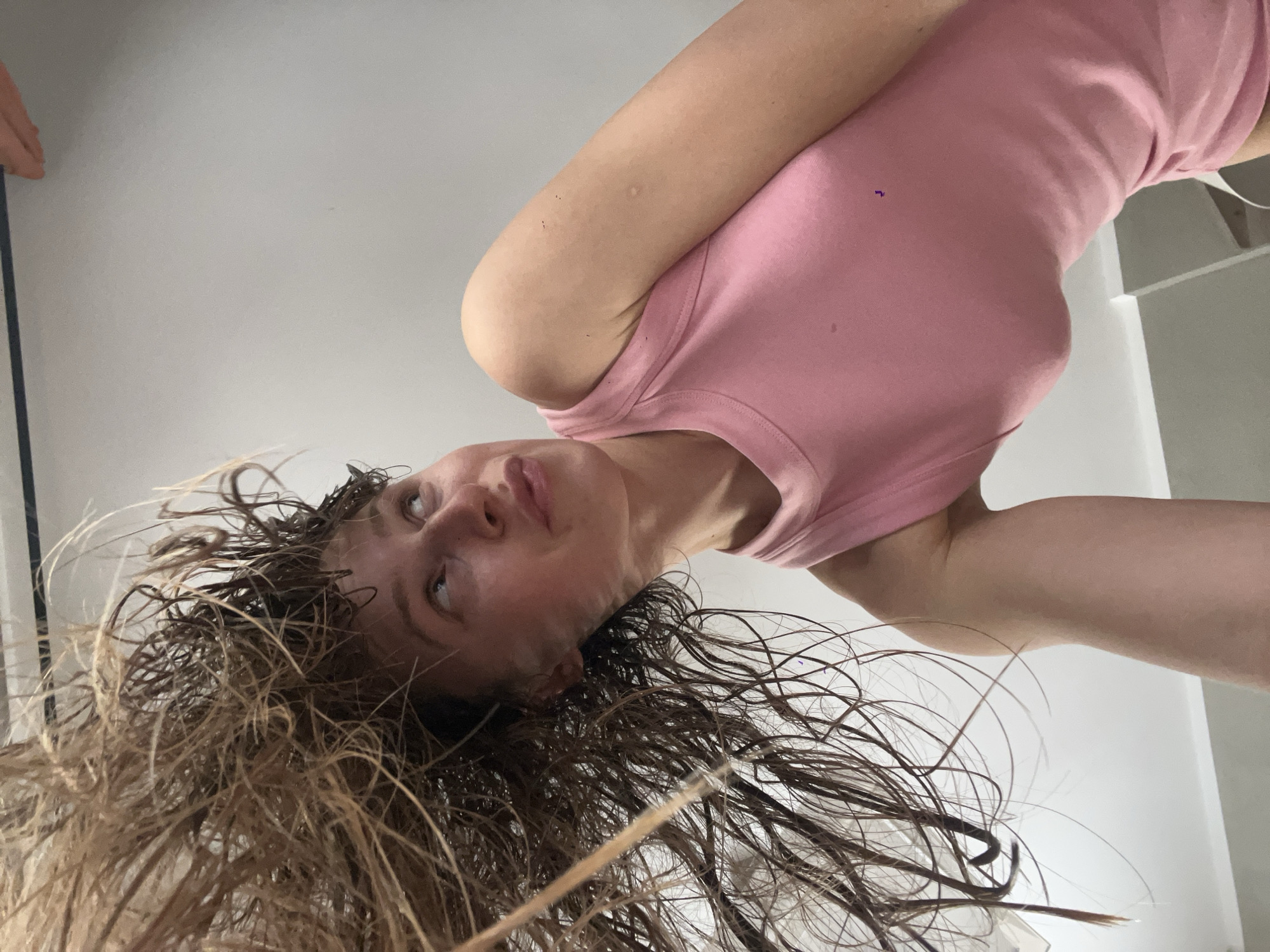 A woman with wet, dark-blonde hair and wearing a pink tank top is seen from a low, inverted angle. Her head is tilted towards the bottom of the frame, and her gaze is directed upwards. Her wet hair is spread around her head and towards the bottom of the image. A white wall is visible in the background. This is likely taken indoors, possibly in a bathroom. There is no explicit indication that this is in Bečići, Montenegro.