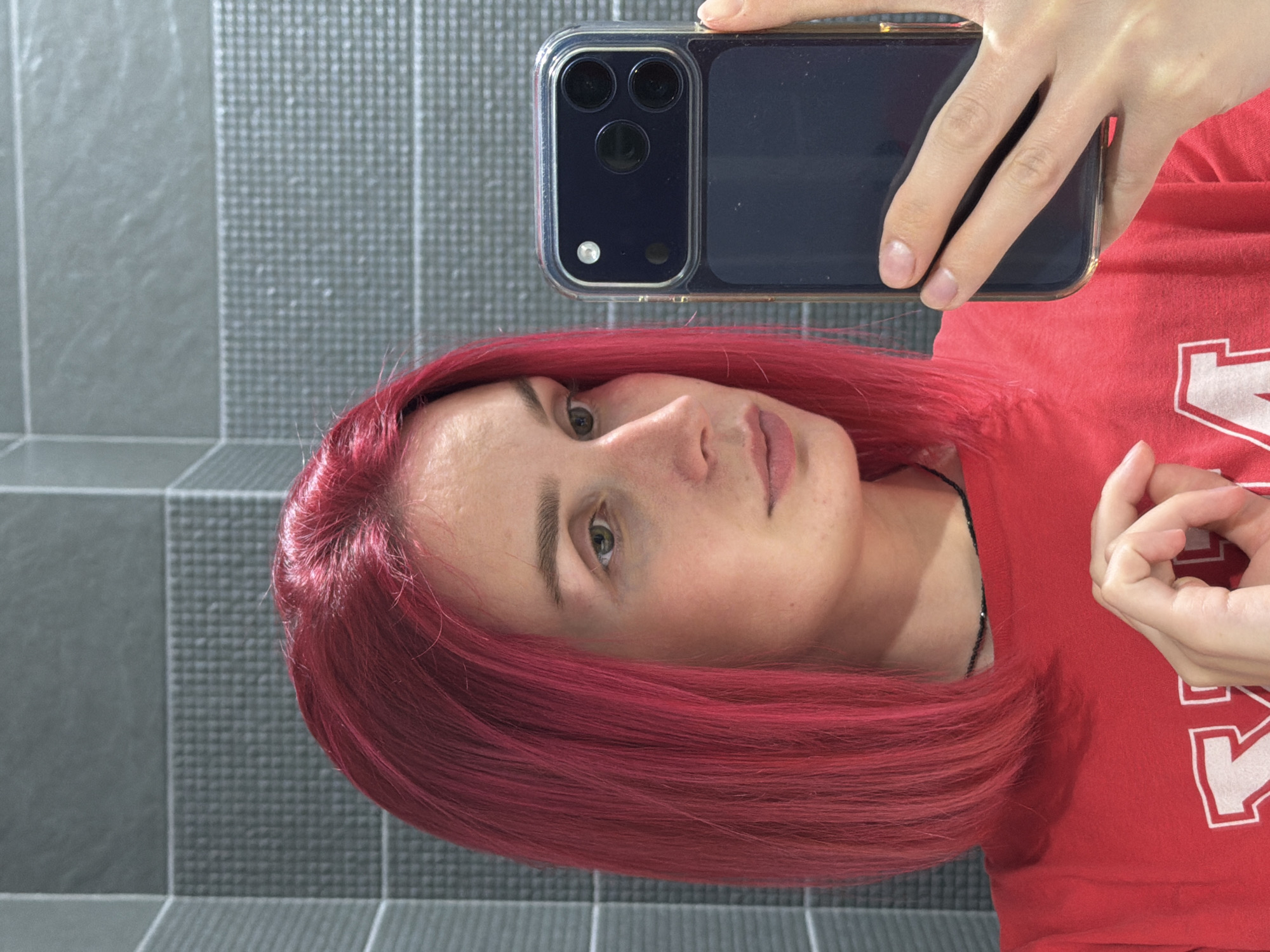 In a well-lit indoor setting with grey tiled walls, a young woman with vibrant pink hair is captured in a selfie. She is wearing a red t-shirt and holding a dark-colored smartphone in her right hand, angled to take the photo. Her left hand is held up near her chest, with her fingers forming a circular shape. The background appears to be a bathroom or similar tiled area. The image does not provide any information to determine its location in the city of Chaikovsky, Russia.