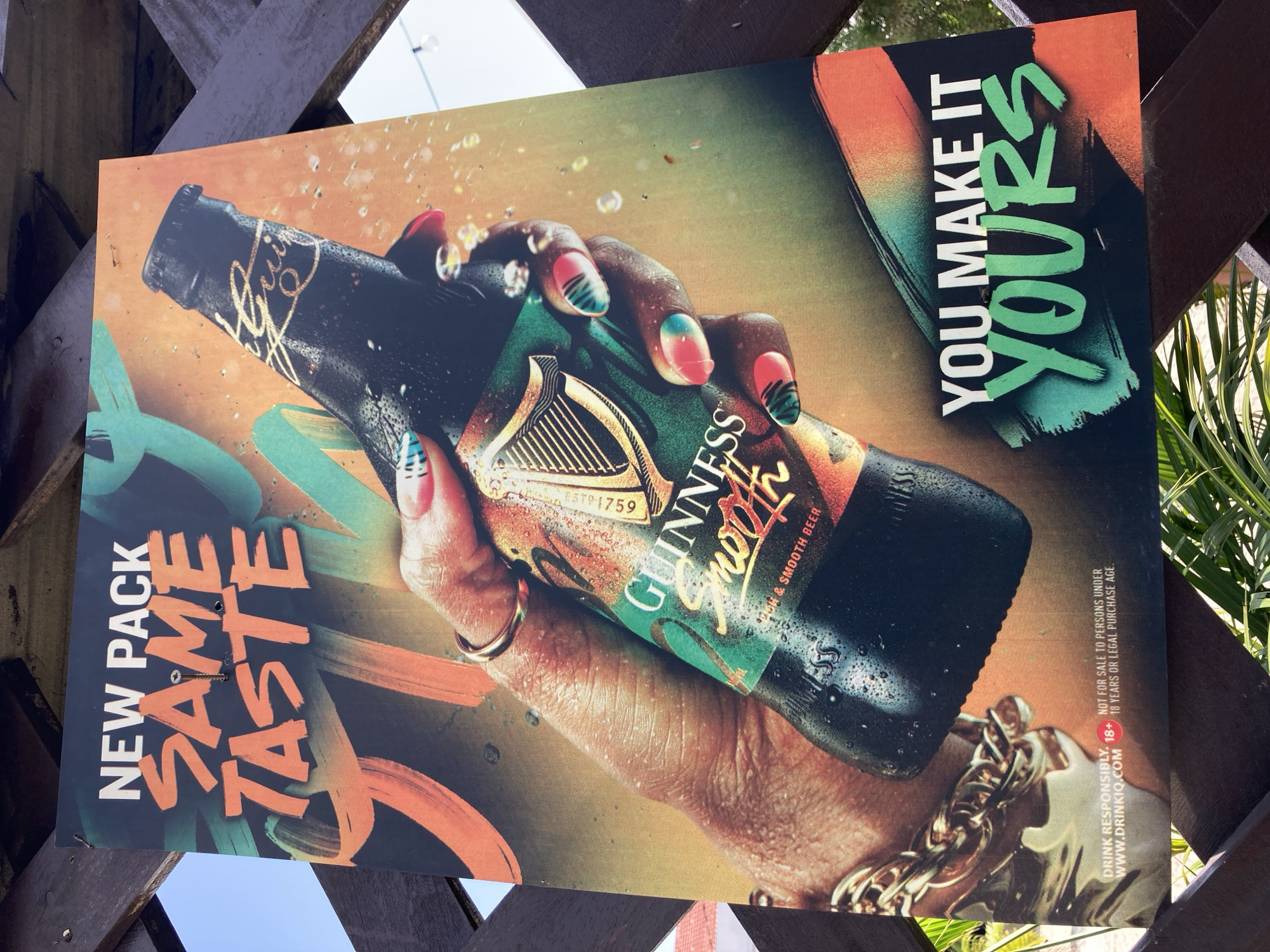 A Guinness beer advertisement is displayed outdoors, mounted on a wooden lattice structure. The ad features a hand holding a sweating bottle of Guinness, with the text "NEW PACK SAME TASTE" prominently displayed at the top in bold orange letters. Below the bottle, the text "YOU MAKE IT YOURS" is written in white and green. The bottom left corner of the ad includes a disclaimer stating "DRINK RESPONSIBLY. 18+ NOT FOR SALE TO PERSONS UNDER 18 YEARS OR LEGAL PURCHASE AGE. WWW.DRINKIQ.COM". The advertisement is positioned outdoors, likely in a public area given its mounting and the background showing foliage and a bright sky. The location in Gwarinpa, Nigeria, is inferred from the context provided.