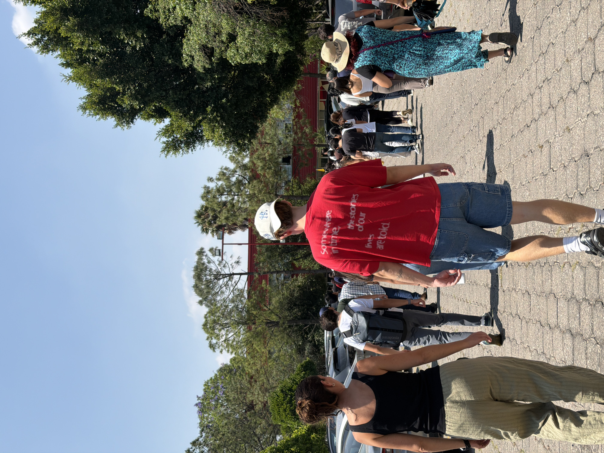 A line of people is standing on a paved surface in San Bernardino Tlaxcalancingo, Mexico. A red building is visible in the background. The sky is blue with some trees on the sides. The people are standing, and some are interacting.