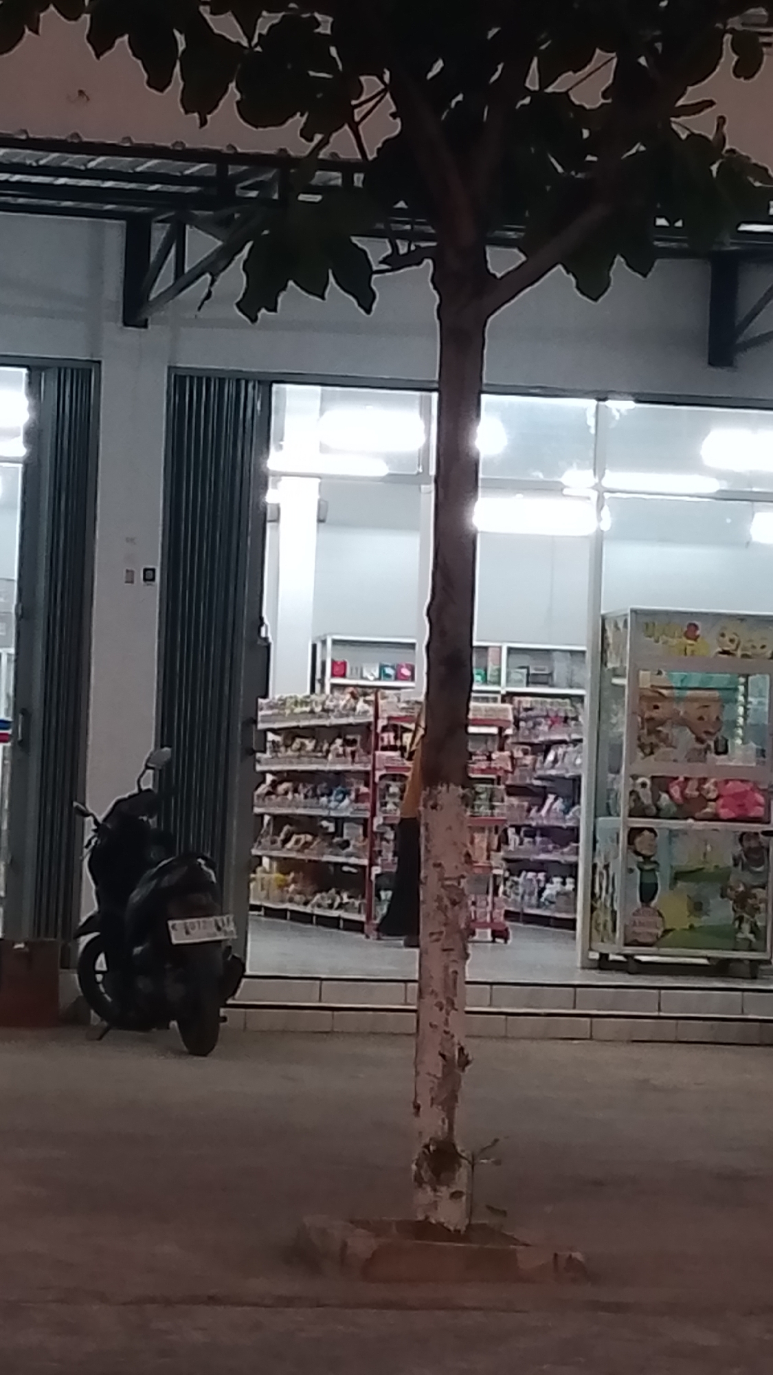 The image depicts the exterior of a convenience store at night in Karanglangu, Indonesia. A tree stands in the foreground, its trunk painted with white markings, with its leaves partially obscuring the building's facade. The store has large glass windows and closed metal roll-up doors. Inside, shelves are stocked with various products, and fluorescent lights illuminate the interior. To the left of the store entrance, a parked motorcycle is visible, its license plate readable as "G 0121 N." The pavement in front of the store appears worn.