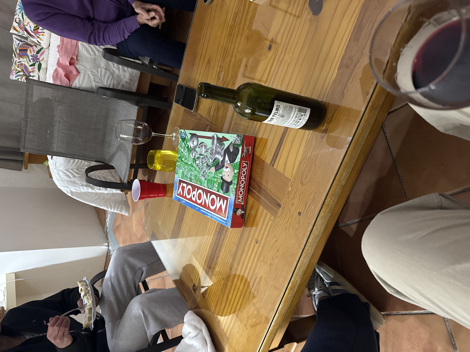 The image depicts a social gathering, possibly a game night, taking place indoors. A wooden table is the central focus, on which a Monopoly game box sits alongside an empty wine bottle, a glass of yellow liquid, a red plastic cup, and a wine glass filled with red wine. Around the table, individuals are seated, with portions of their attire visible. One person wears a purple sweater, another has on grey sweatpants, and another is wearing beige pants and grey sneakers. Someone is holding a plate of food and eating with a fork. The location is indicated as Puebla, Mexico, but the specifics of the room or immediate surroundings are not clear from the image. The actions are primarily focused on eating and social interaction around the table, presumably engaged in a game.