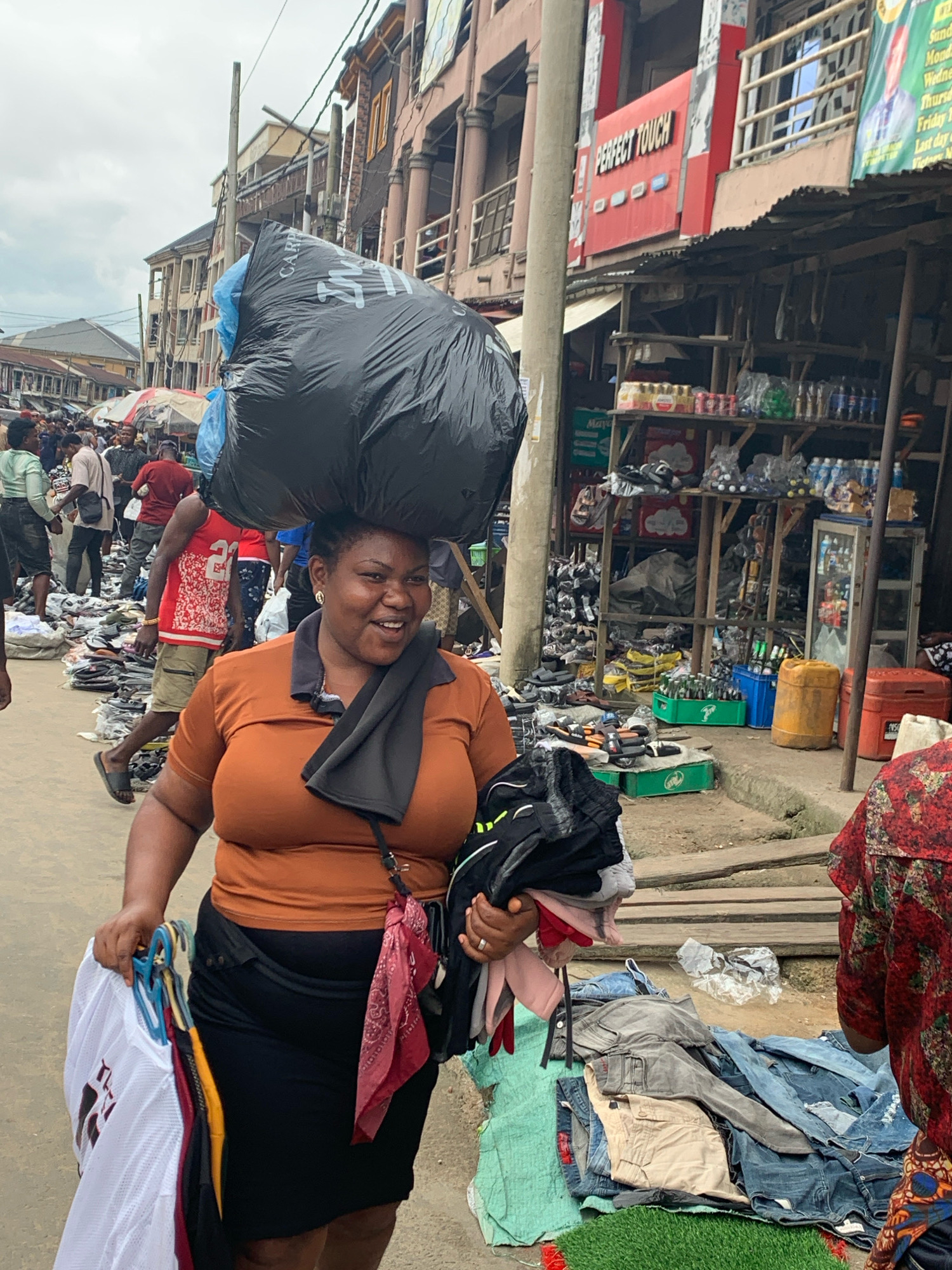 A woman carries a large black plastic bag filled with goods on her head in a crowded market scene in Aba, Nigeria. She is smiling and holding several items of clothing on hangers in her left hand. The woman is wearing an orange top and a black skirt. In the background, other people are seen walking and shopping, with stalls and shops lining the street. The ground is paved, and there are various goods displayed for sale, including clothing, shoes, and drinks. The overall environment suggests a busy and lively marketplace.