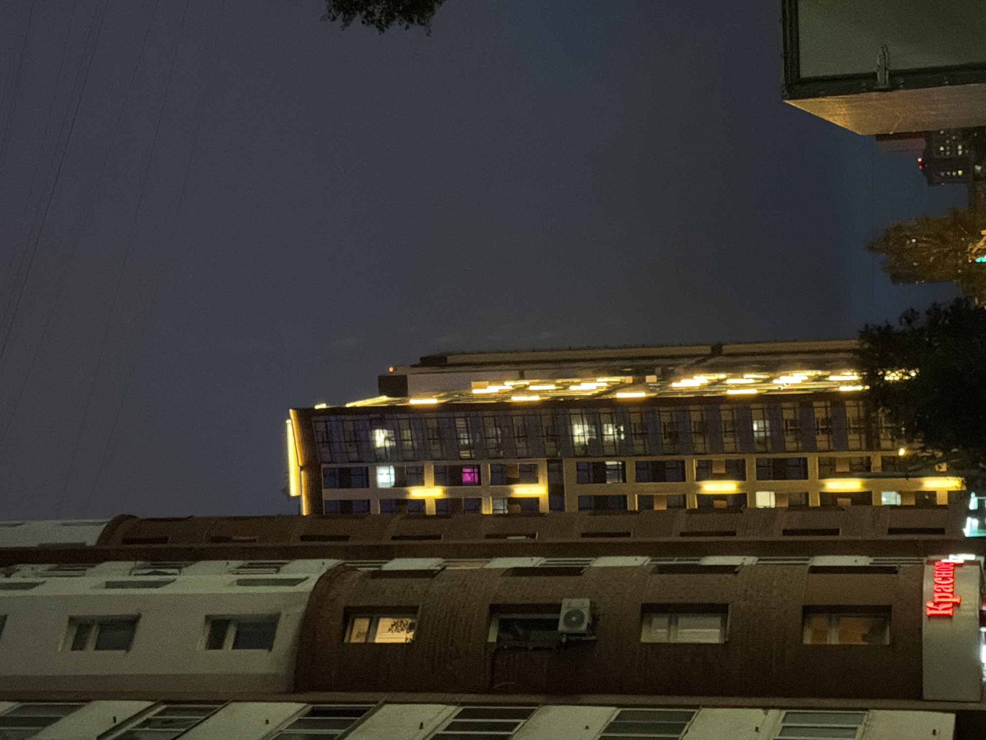 The image shows a nighttime view of apartment buildings in Yekaterinburg, Russia. The buildings are illuminated by interior lights and exterior decorative lighting. On the left, a multi-story apartment building with a curved brick facade is visible, with several windows lit and an air conditioning unit mounted on one of the lower floors. Below this, a red neon sign that reads "Красно" is partially visible, likely indicating a shop or business. To the right of this building is another, taller apartment building with a modern design, featuring a facade of glass and vertical yellow lights. Several windows on this building are also lit, with some displaying pink light. In the background, more buildings are visible in the distance, some with scattered lights. The sky is dark and overcast, with power lines crossing the upper portion of the frame. Trees are visible in the lower right corner and also in the distance. The overall impression is of a residential urban environment at night.