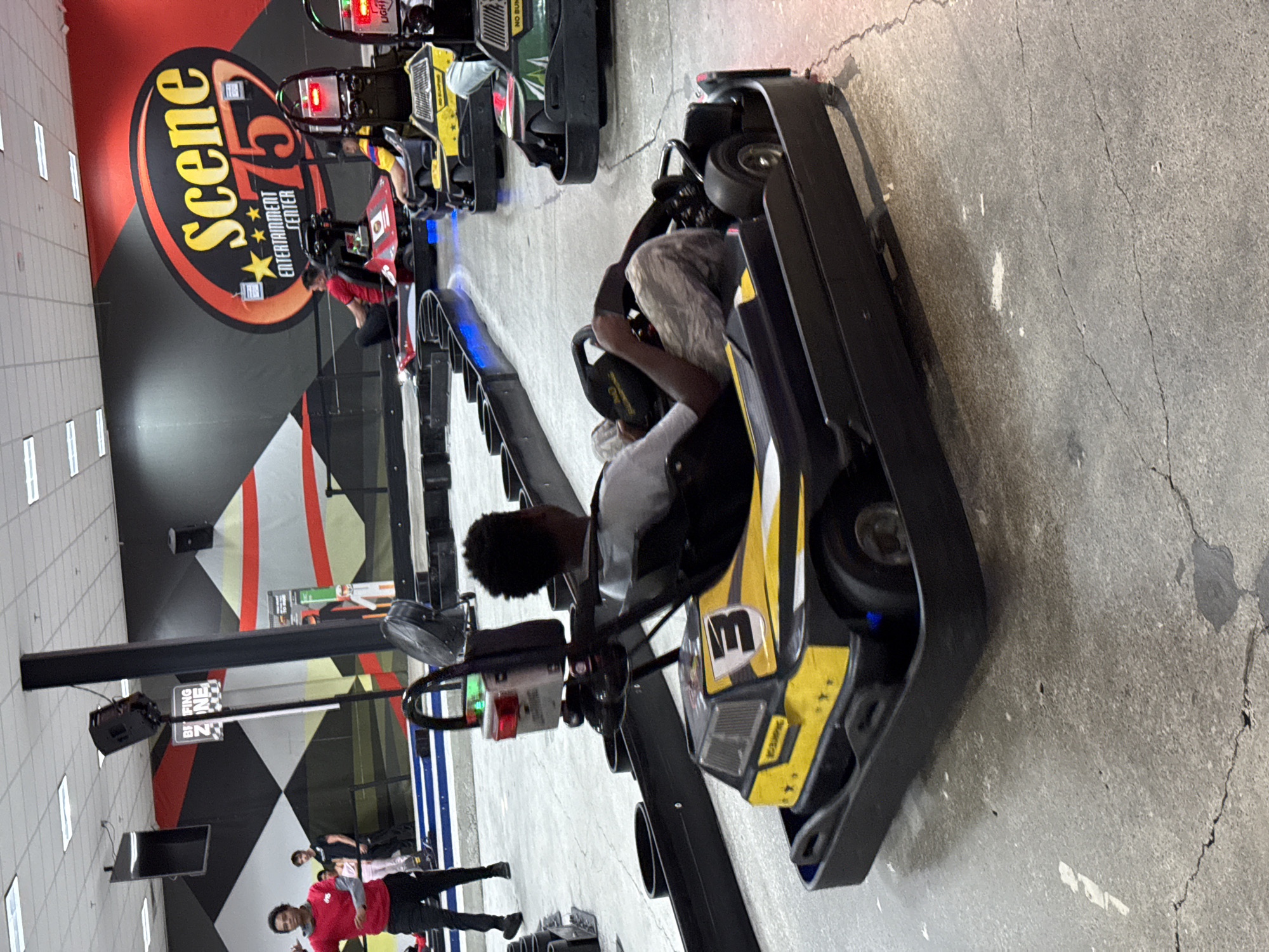 The image shows the interior of Scene75 Entertainment Center, a family entertainment complex in Columbus, Ohio, United States. In the foreground, an individual is sitting in a black and yellow go-kart with the number "13" on it, suggesting a go-kart racing activity. The go-kart is positioned on a concrete surface, showing some wear and tear. In the background, other go-karts are lined up, with additional people visible. The wall behind the go-kart area features a checkered black and white pattern and the logo of Scene75. Above, there are ceiling lights and what appears to be a speaker and a screen mounted on a beam. In the distance, there's a group of people, one wearing a red shirt.