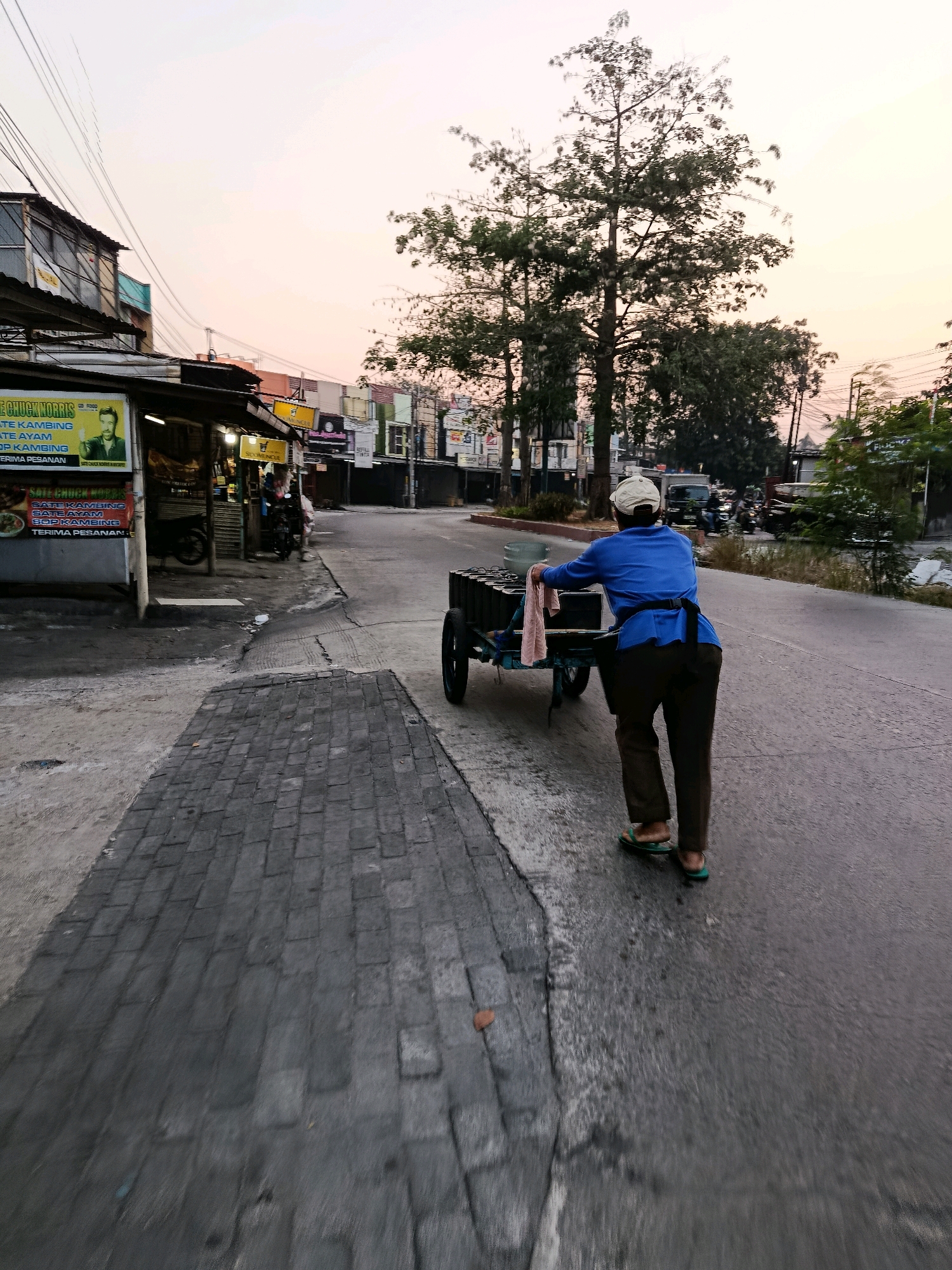 An individual is captured from behind, pushing a cart filled with items down a street in Bekasi, Indonesia. The person is wearing a blue shirt, dark pants, a white cap, and sandals. The cart appears to be loaded with multiple stacked objects, possibly containers or supplies. The street has a paved sidewalk with cobblestone-like patterns on the left side, and the road surface shows signs of wear and tear. Shops and buildings line the street, some with signage, suggesting a commercial or residential area. Several trees are visible, and the sky exhibits the soft hues of either sunrise or sunset. The overall scene suggests an everyday urban activity.