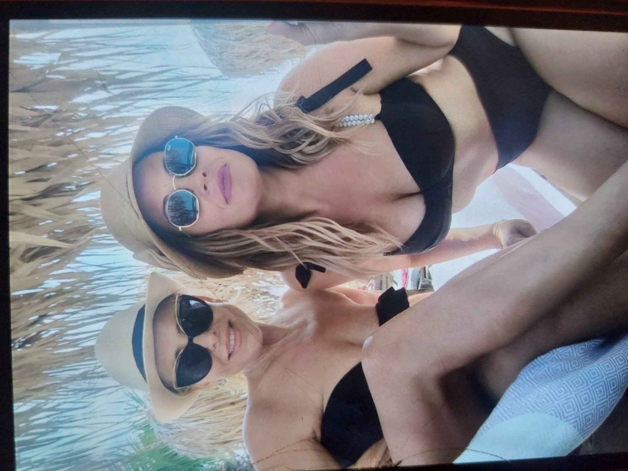 The image displays two women relaxing outdoors, likely at a beach or resort in Tsarevo, Bulgaria, under a thatched roof or umbrella providing shade. The photograph is taken from a low angle, looking upwards at the subjects who appear to be lying down.
The woman in the upper portion of the frame has long, wavy blonde or light brown hair. She is wearing a light brown straw hat with a dark band and round, reflective sunglasses. Her lips are colored with pink lipstick, and she has a subtle, relaxed smile. She wears a black bikini top with a strap featuring small white beaded or pearl-like embellishments.
The second woman, positioned in the lower half of the frame, also has blonde or light brown hair and wears a similar light brown straw hat with a dark band. She is wearing dark, cat-eye style sunglasses and exhibits a broad, cheerful smile, revealing her teeth. She is dressed in a black bikini top, and her legs are visible, with one resting on a light blue and white patterned towel or blanket.
The background shows a bright, light blue sky and some light-colored foliage, suggesting a sunny day. Both women appear happy and relaxed, enjoying their time in the coastal setting of Tsarevo. There is no visible text in the image.
