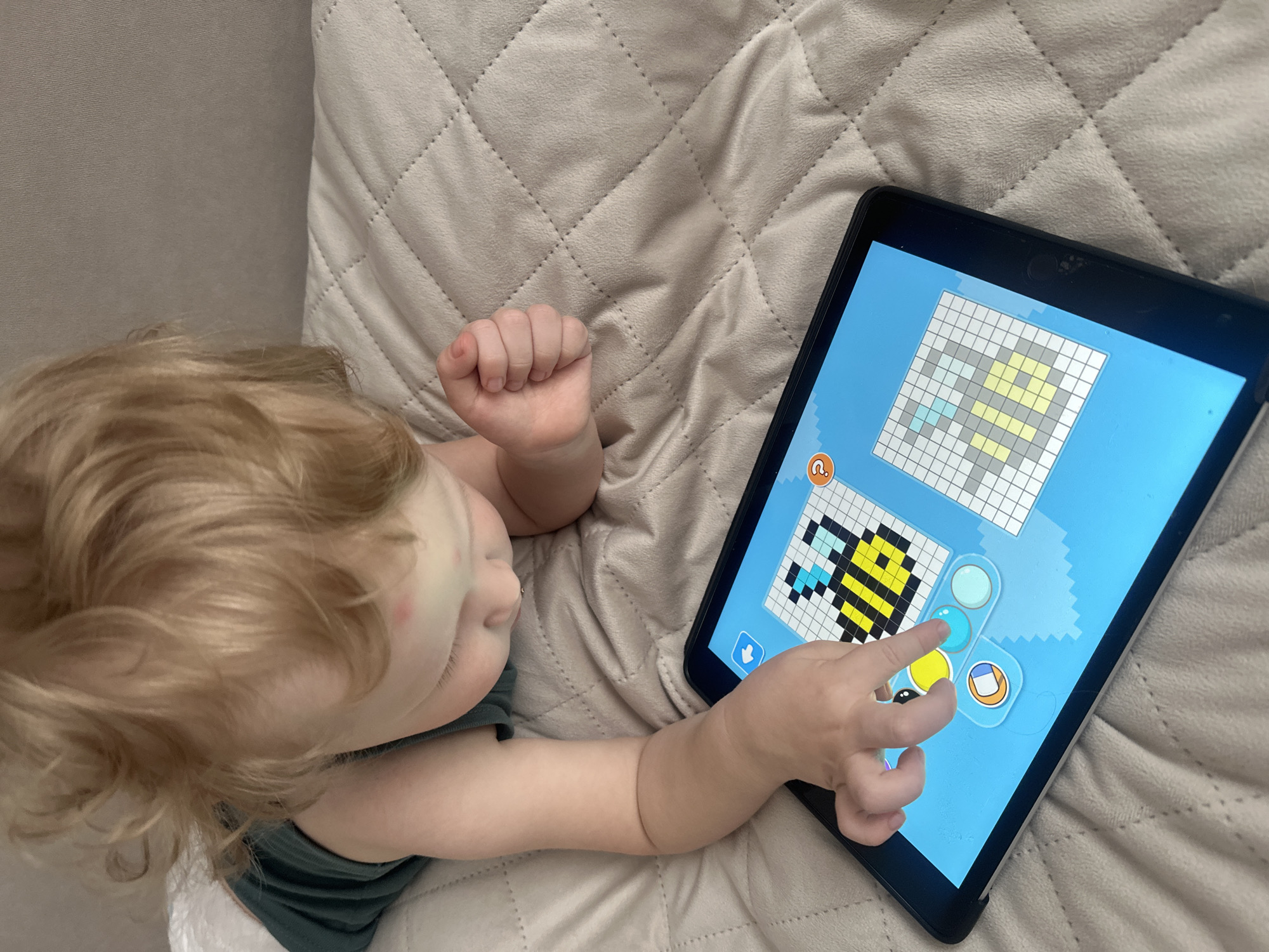 A young child is lying on a light beige quilted surface, possibly a bed or sofa, playing a pixel art game on a black tablet. The tablet screen shows a partially completed image, with a bee pixel art design in the lower-left corner. The child is pointing with their right index finger at the screen. The environment is simple and suggests a domestic setting. The photo does not have information to determine if it was taken in Votkinsk, Russia.